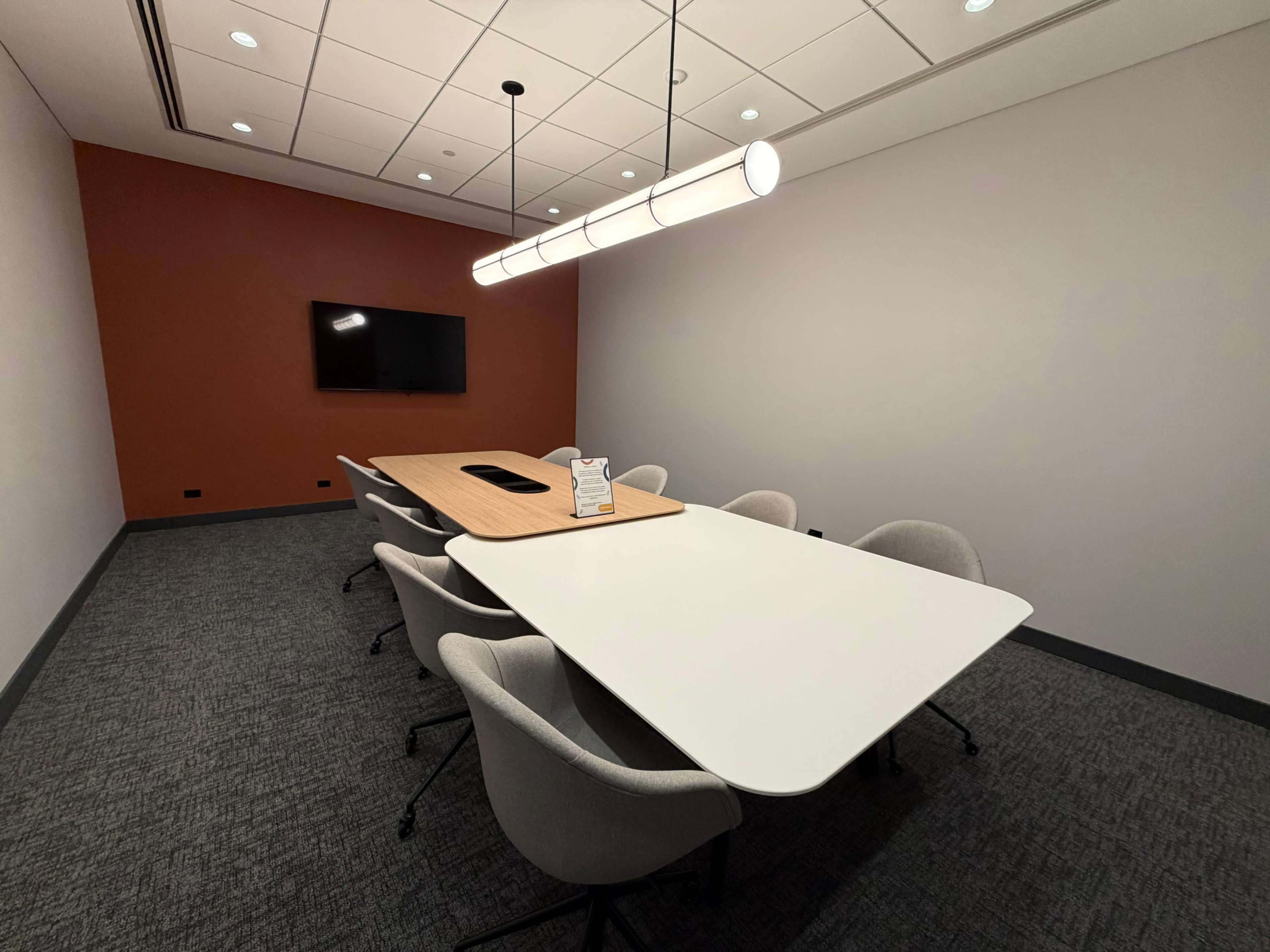 The image shows a modern conference room with a large table, several chairs, a television mounted on the wall, and a pendant light overhead.