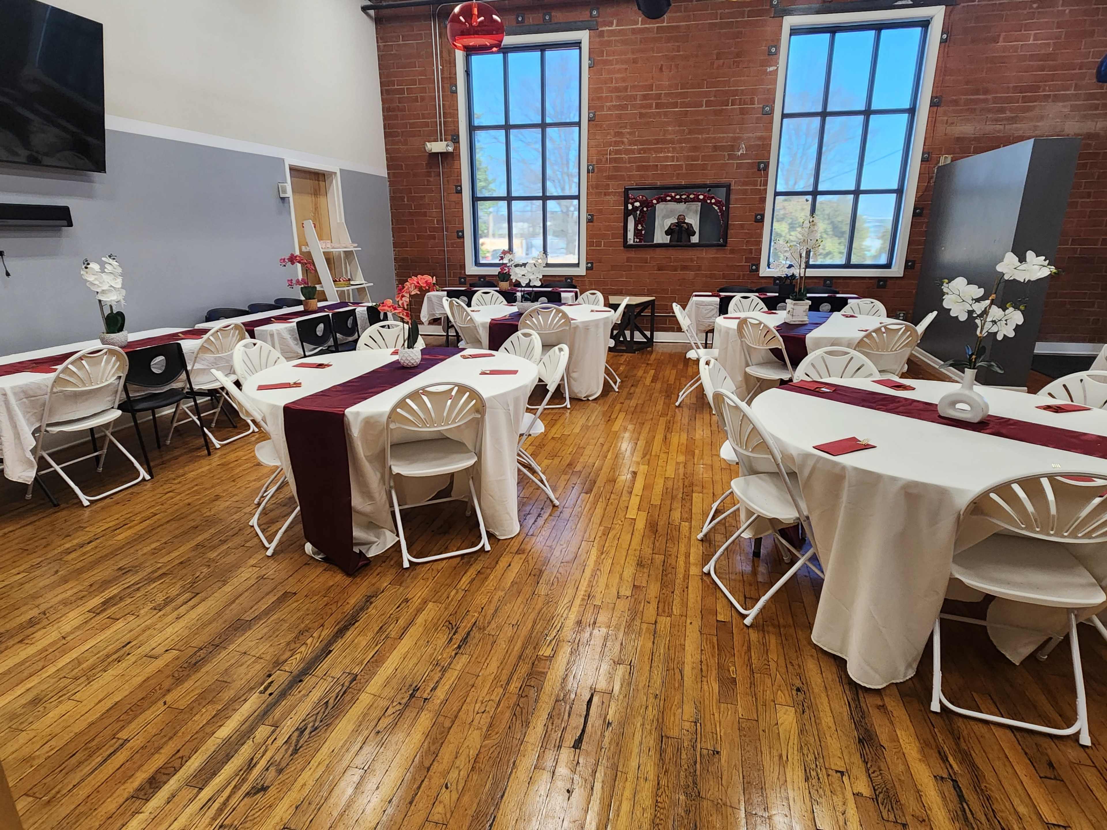 The image shows a spacious interior with several round tables dressed in white tablecloths and burgundy runners, set up for an event in a room with brick walls and large windows.