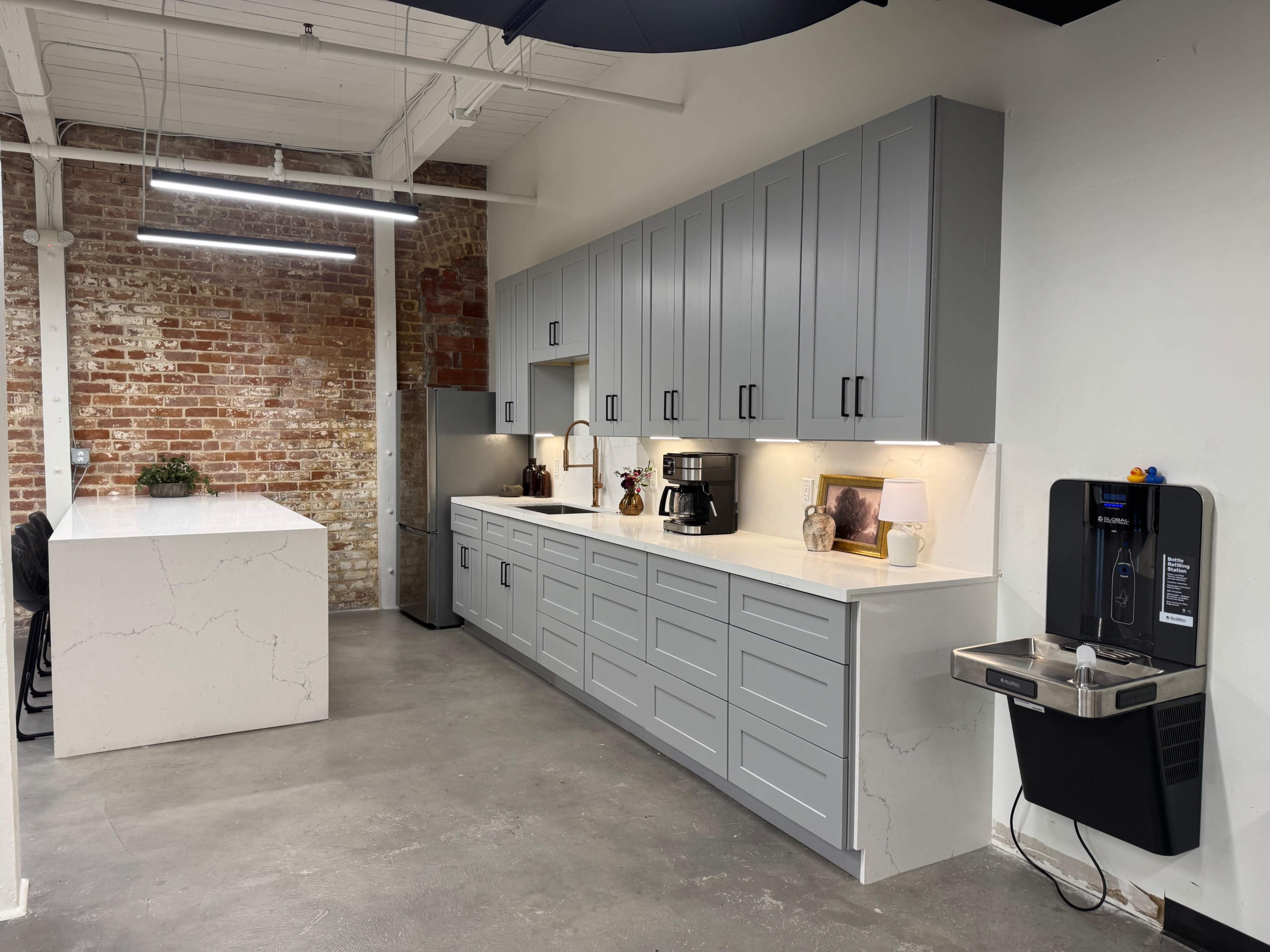 The image shows a modern kitchen area with gray cabinets, a white countertop, a coffee maker, and a small sink against a backdrop of exposed brick walls.