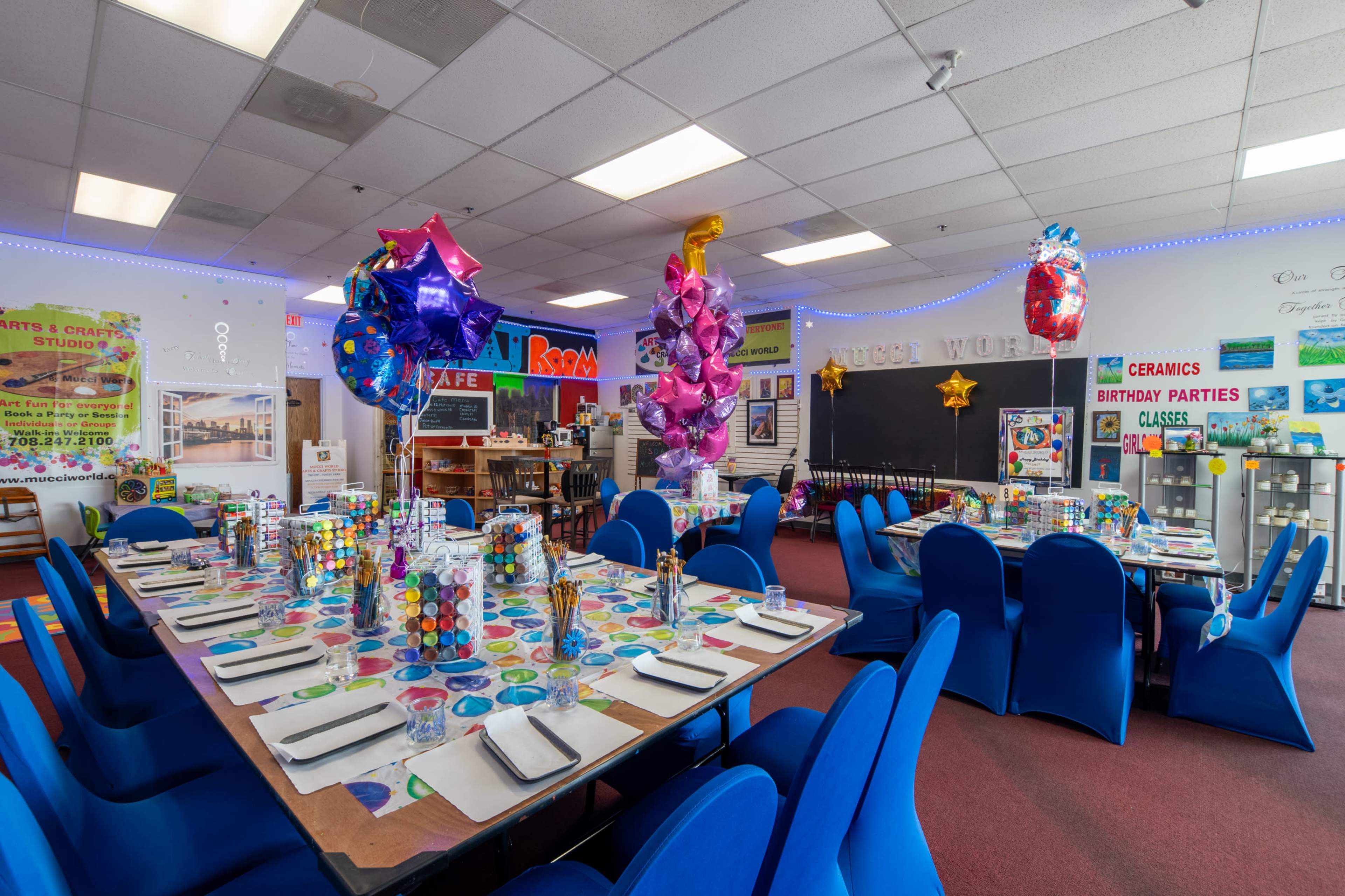 A brightly decorated party space with a long table set for a celebration, featuring colorful tablecloths, centerpieces, and balloons.