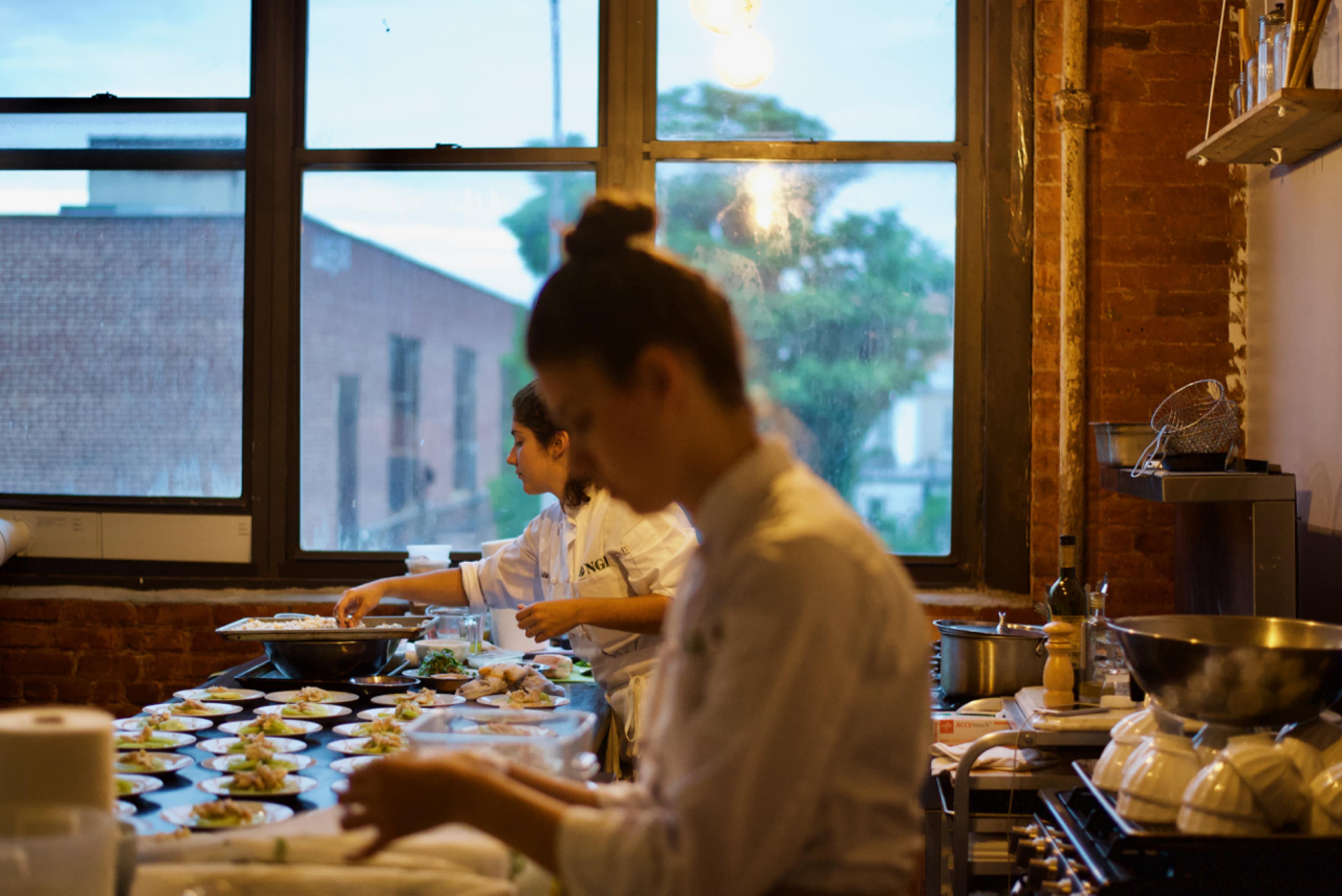 Two chefs are preparing plated dishes in a restaurant kitchen with large windows overlooking an urban landscape.