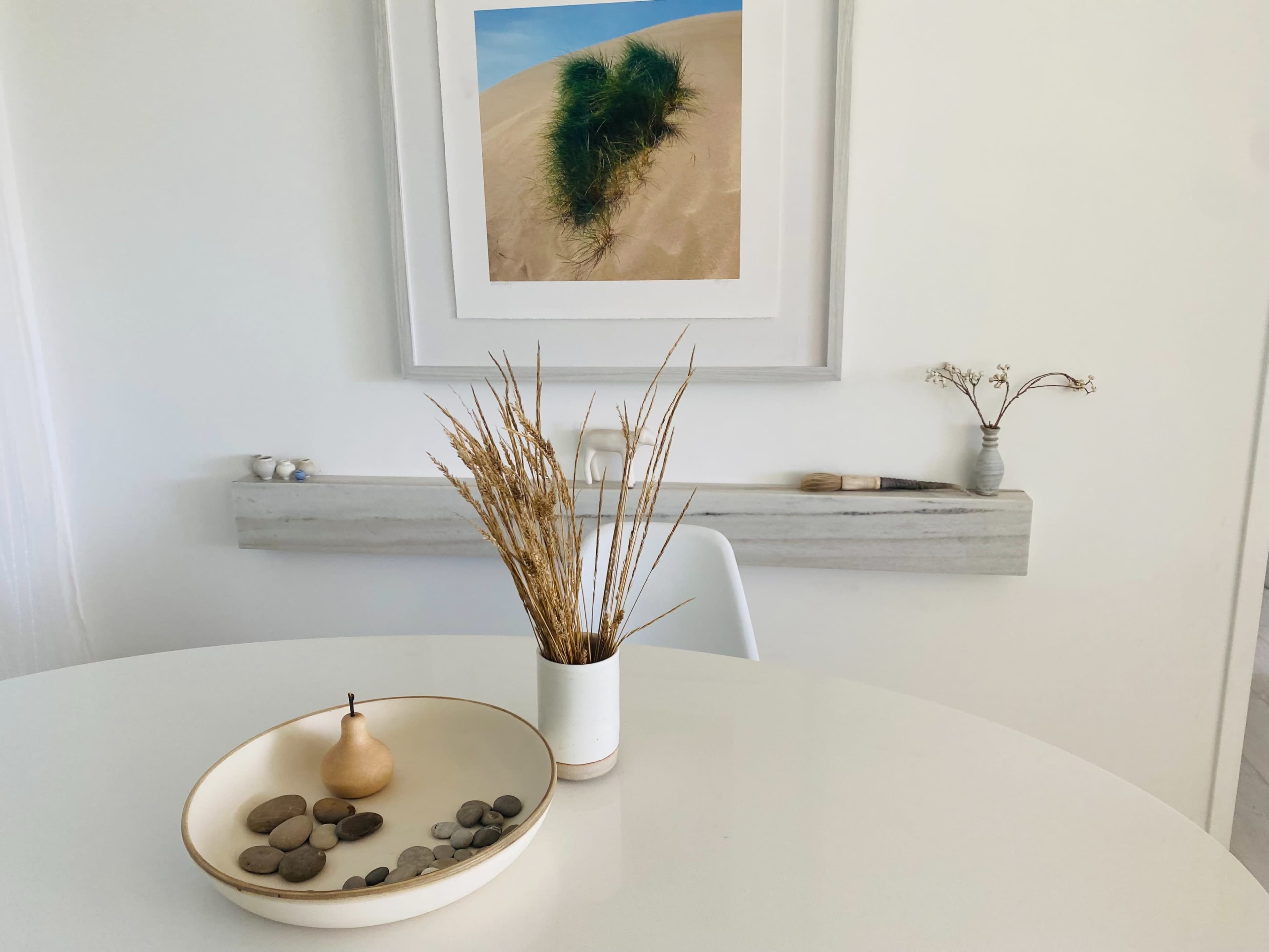 A minimalist dining table features a bowl of stones, a pear-shaped object, and a vase with dried grasses, set against a backdrop of a framed photograph of sand dunes.