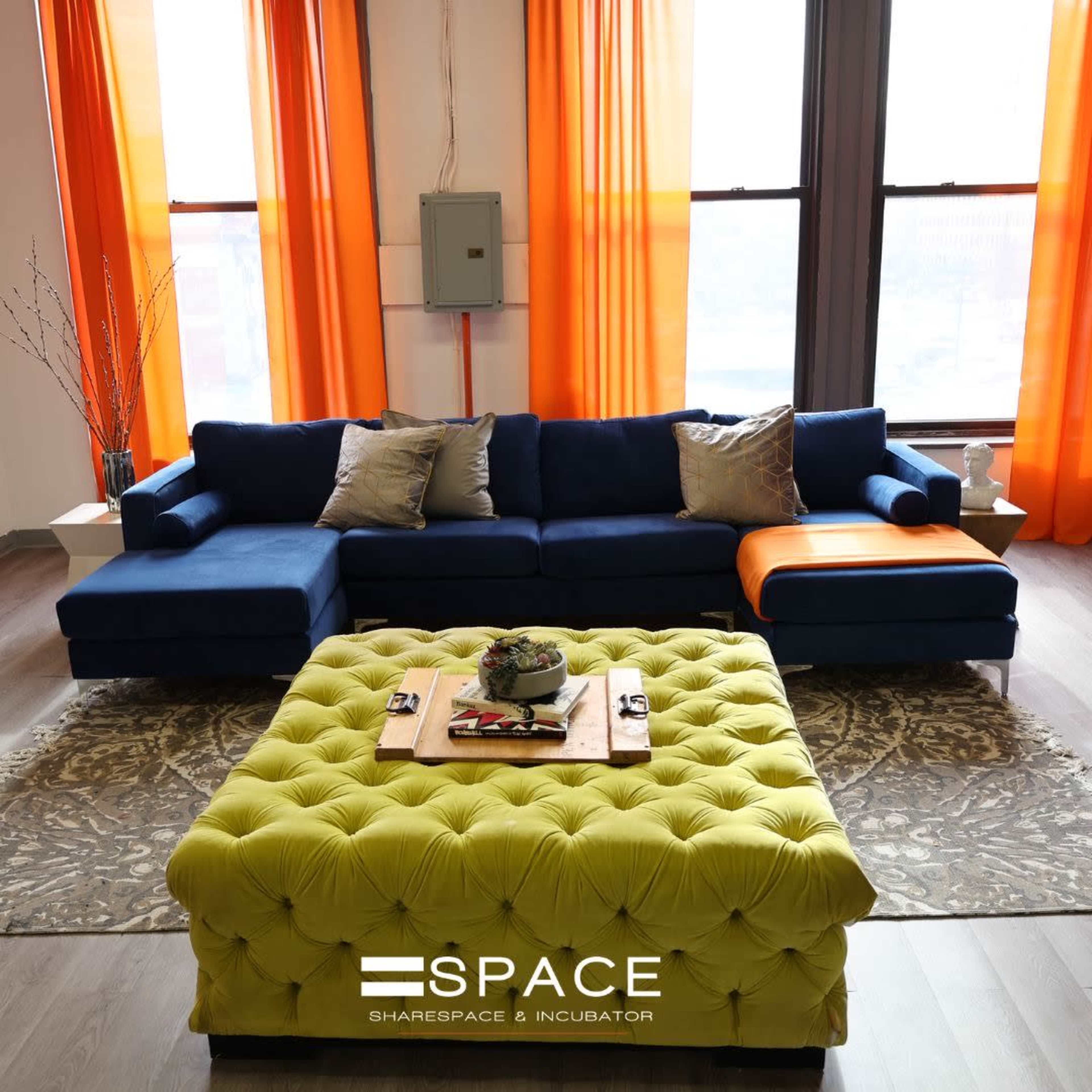 A modern living room features a blue sectional sofa with decorative pillows and a large green tufted ottoman on a patterned rug, complemented by orange curtains.
