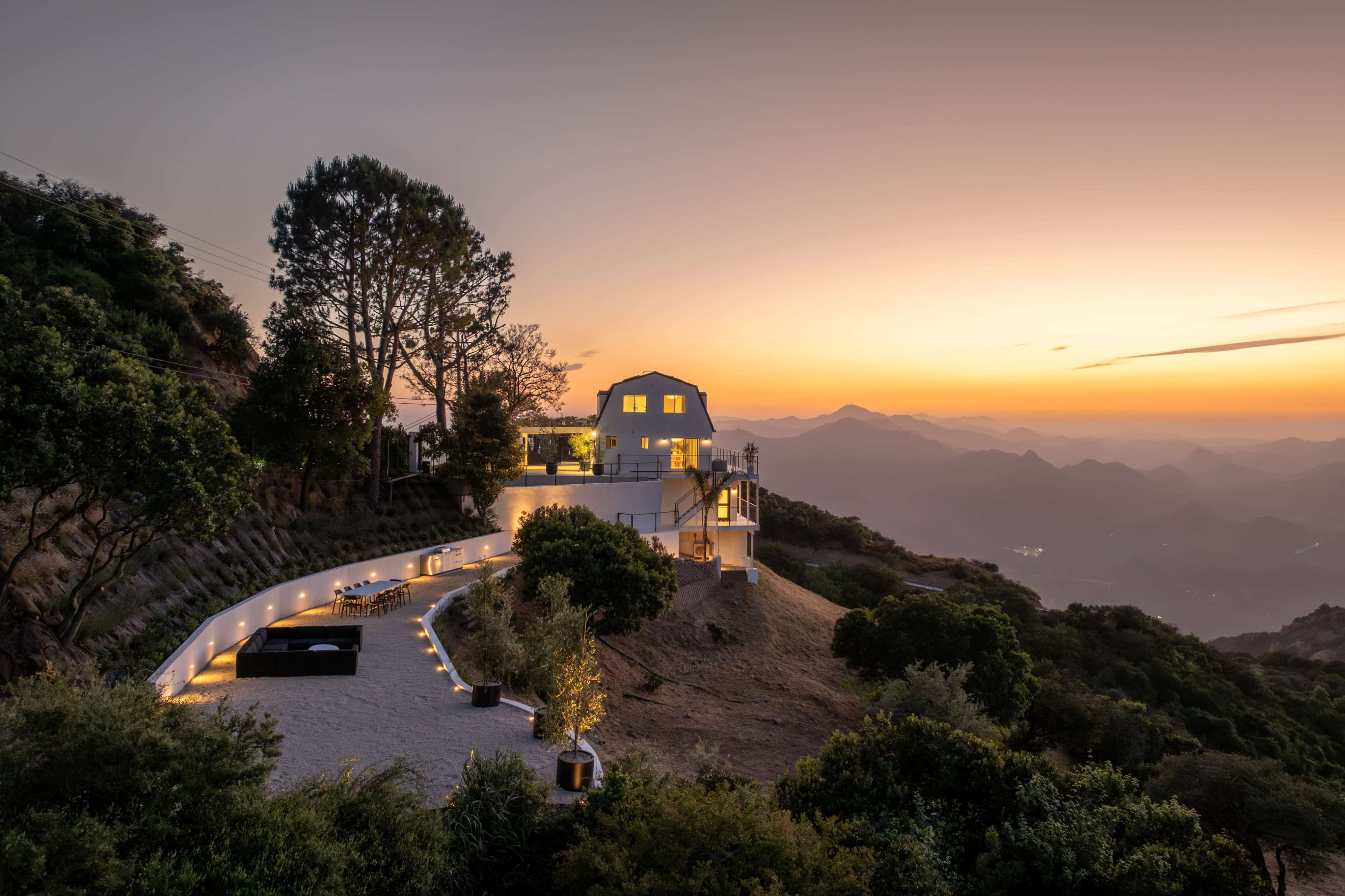 A modern white house is situated on a hillside, overlooking a mountain landscape at sunset with lights lining a paved terrace.