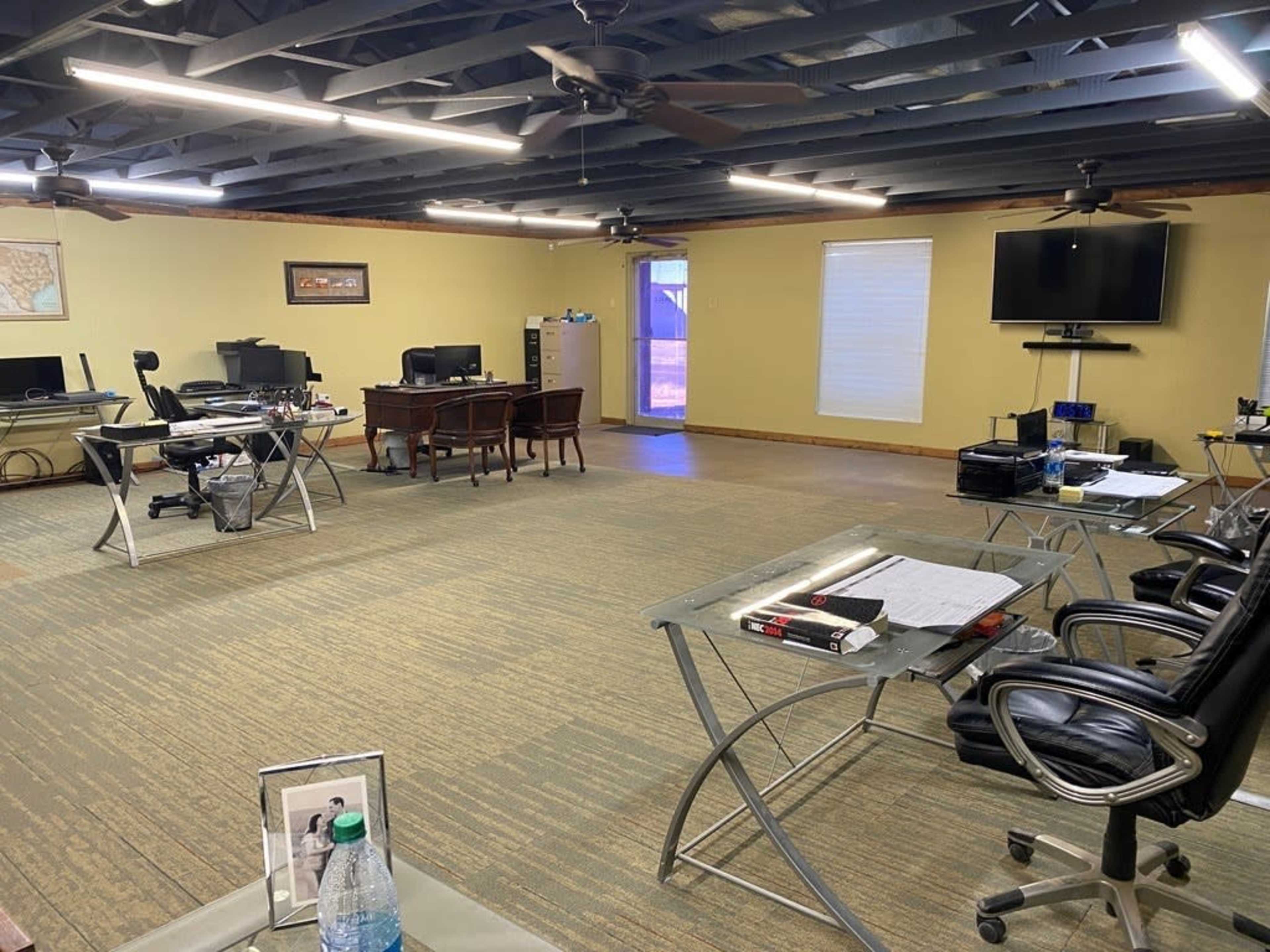 The image shows a spacious office with multiple workstations, including desks and chairs, arranged around a central area, and equipped with computers and a television.