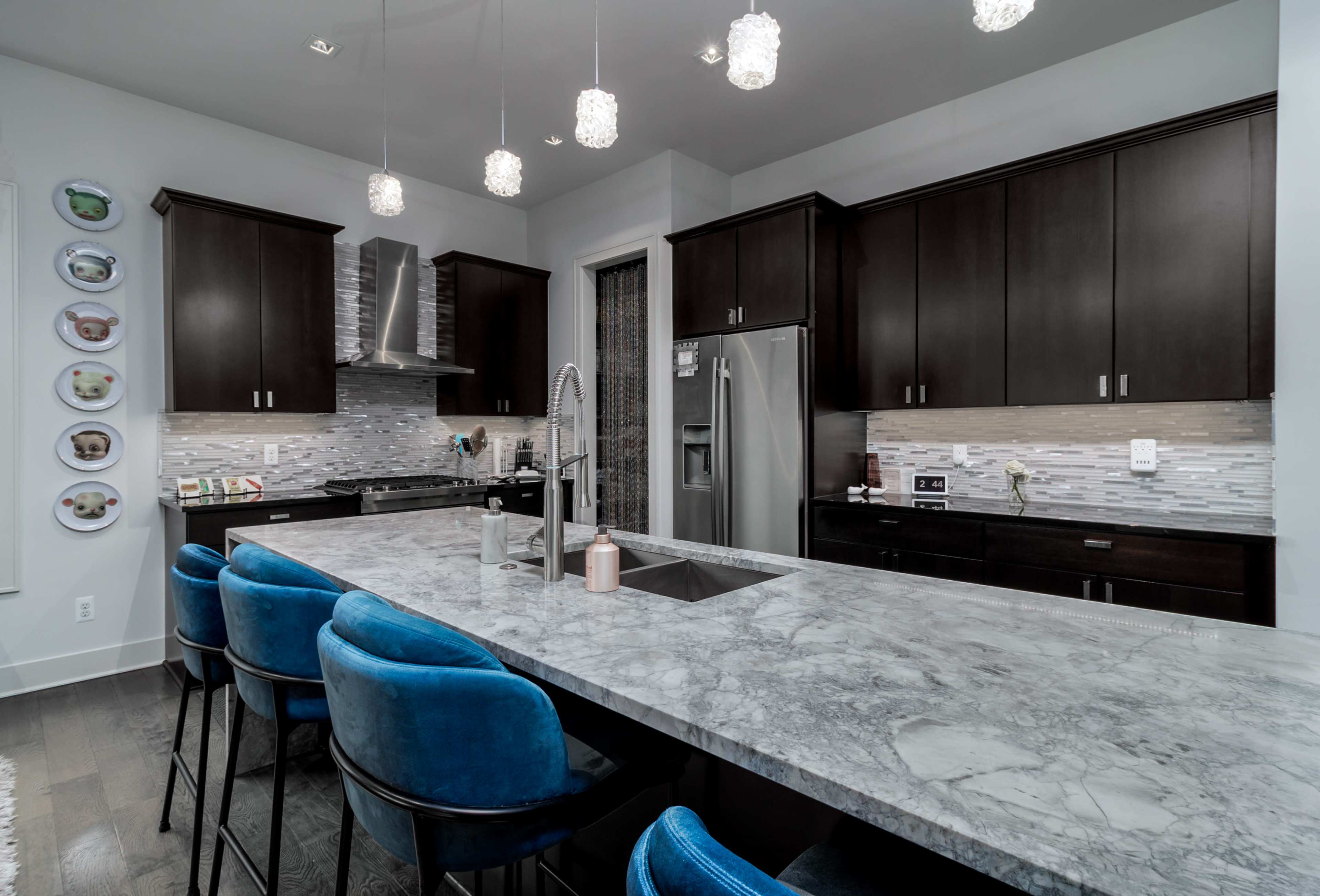 The image shows a modern kitchen featuring dark cabinetry, a marble countertop, and blue upholstered bar stools along a large island.