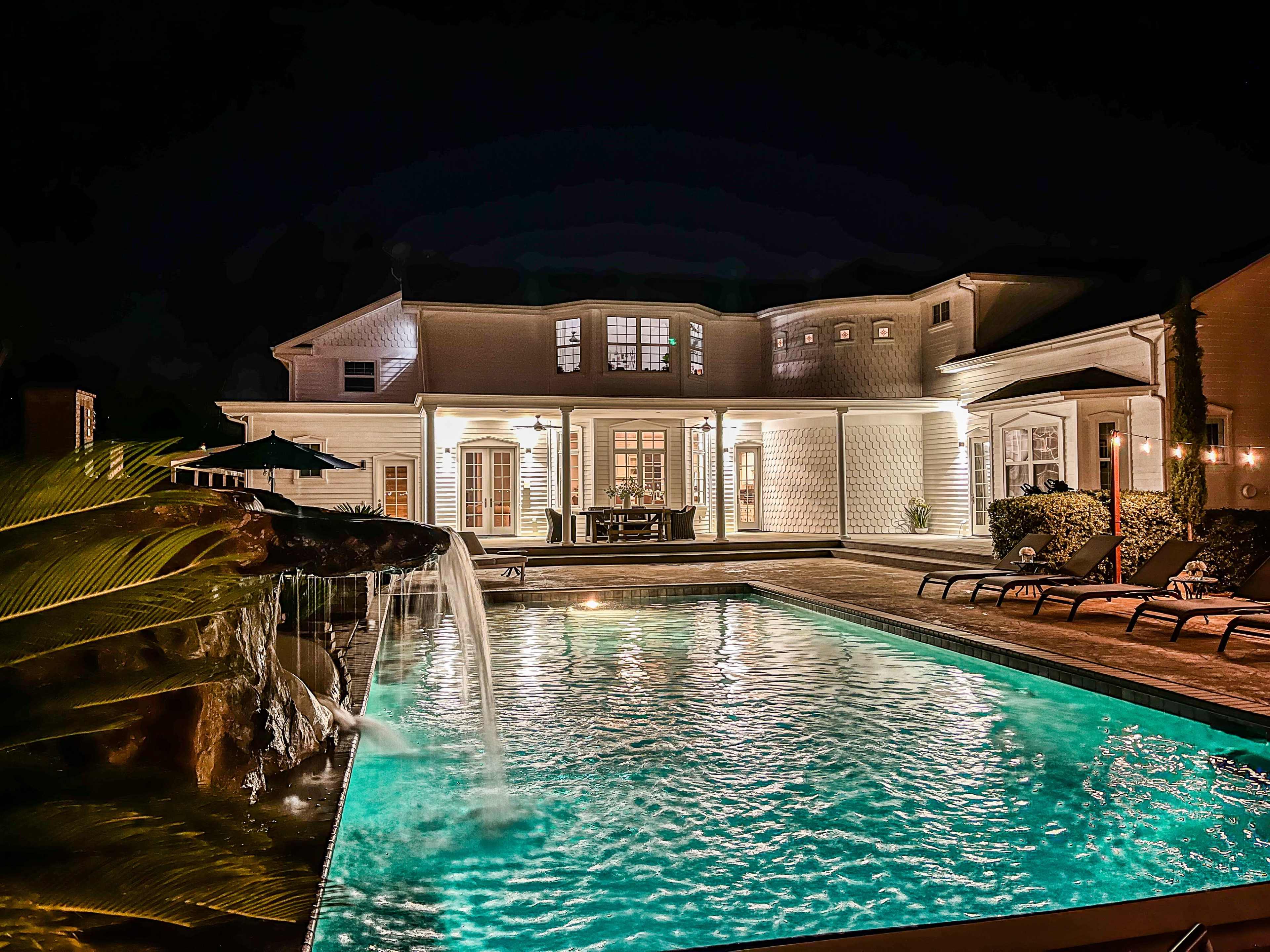 The image shows a well-lit house at night with a pool and a waterfall feature, surrounded by lounge chairs and patio furniture.
