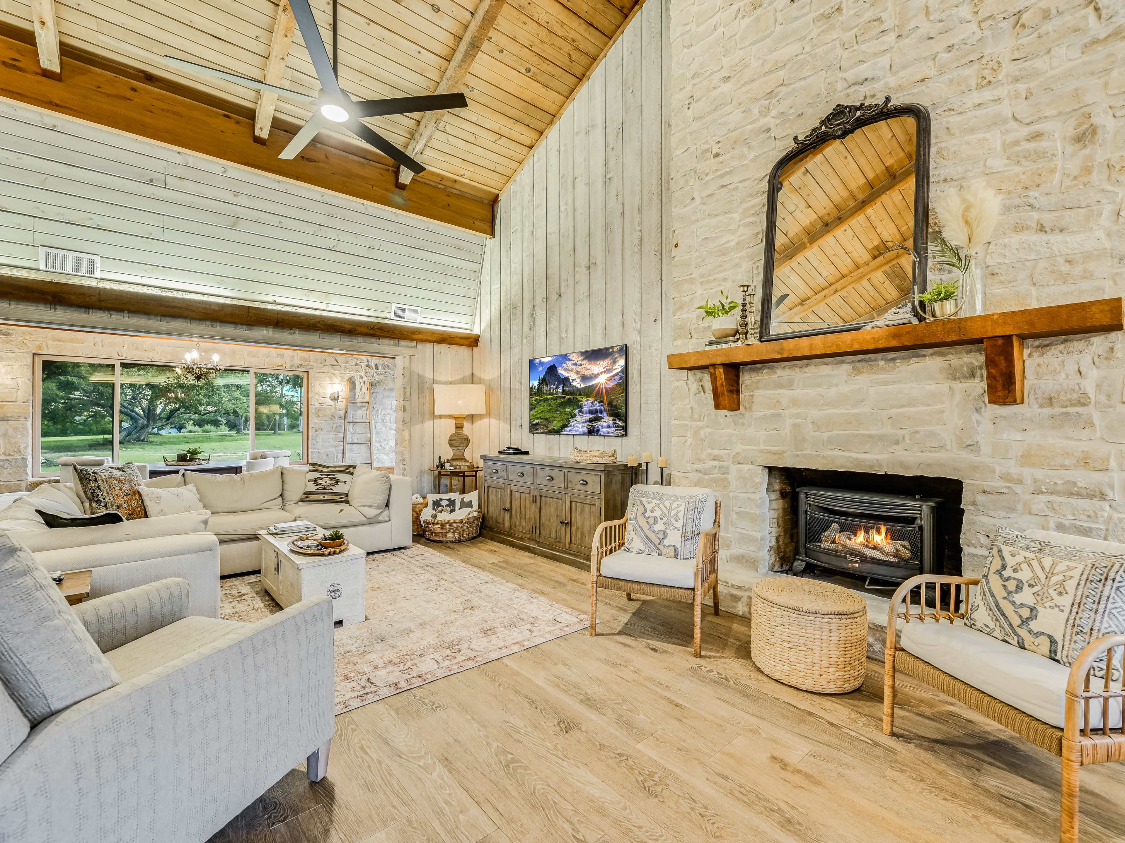 A spacious living room with a stone fireplace, wooden beams, and large windows overlooking a green outdoor area.