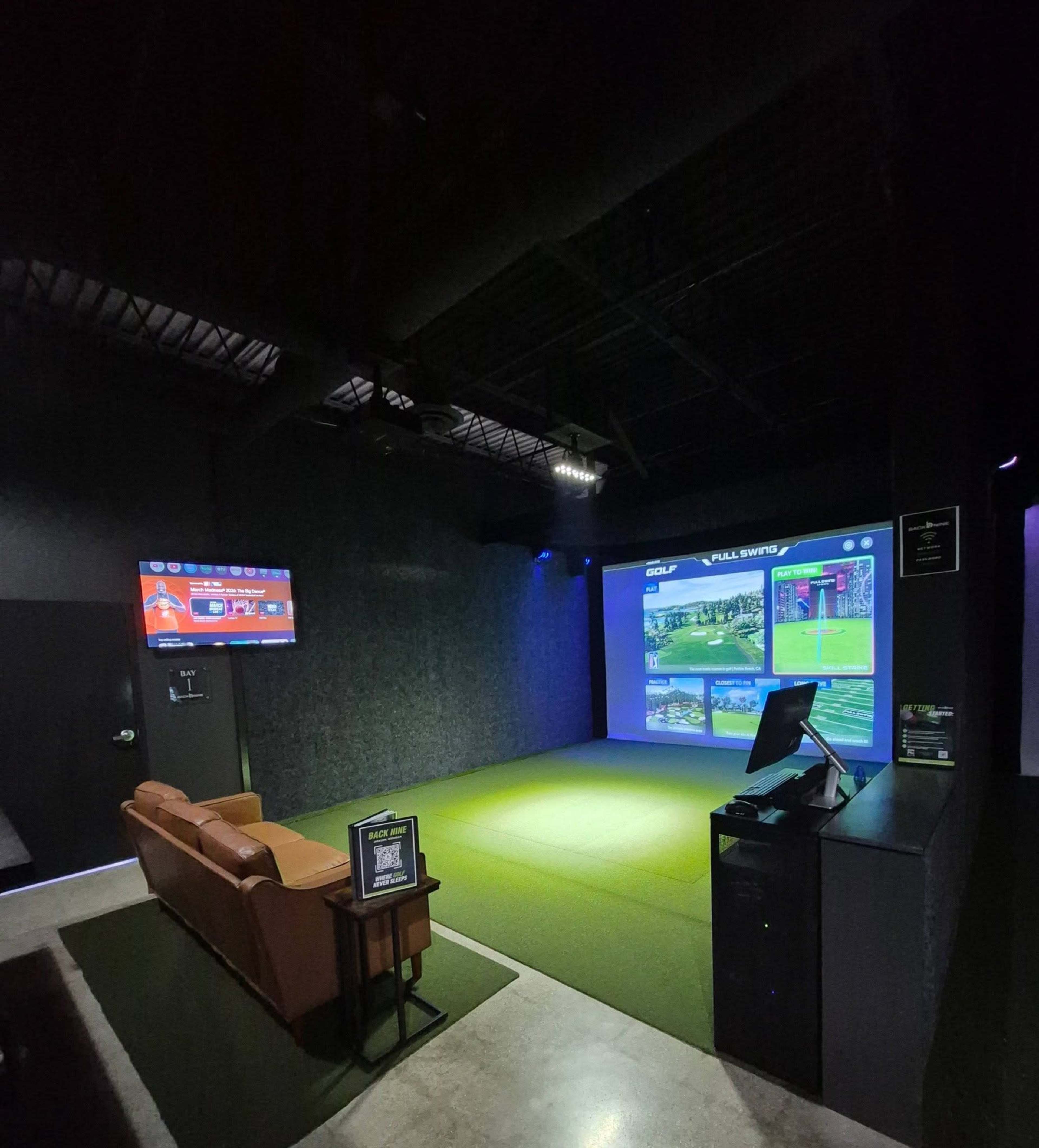 The image shows an indoor golf simulator room featuring a large screen displaying a golf course, a seating area with a brown couch, and a monitor on the wall.
