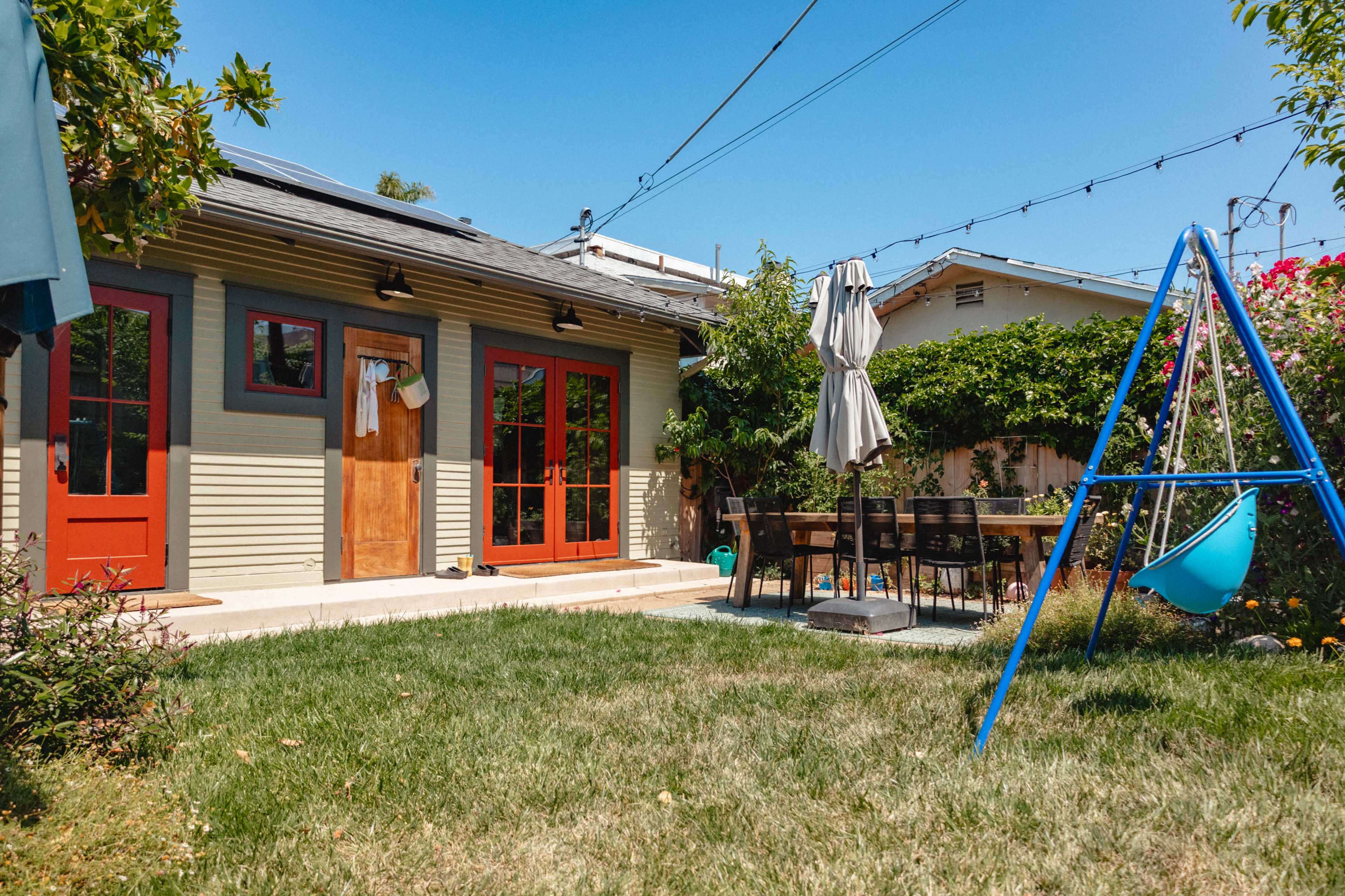 The image shows a backyard with a sliding glass door entrance to a house, a dining table with chairs, a swing set, and lush greenery.