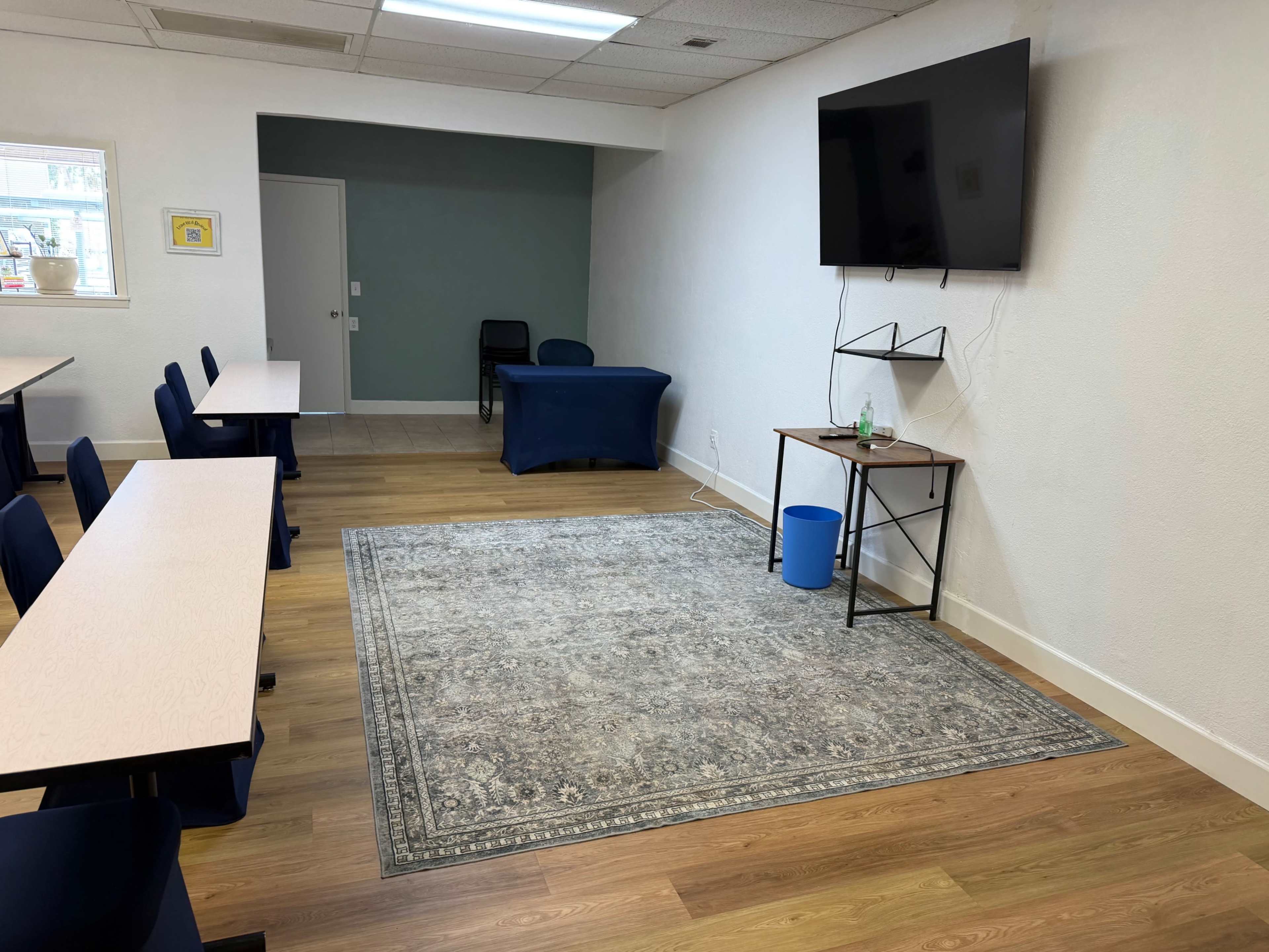 The image shows a sparsely decorated classroom or meeting space with several empty tables and chairs, a television mounted on the wall, and a blue table against one wall.