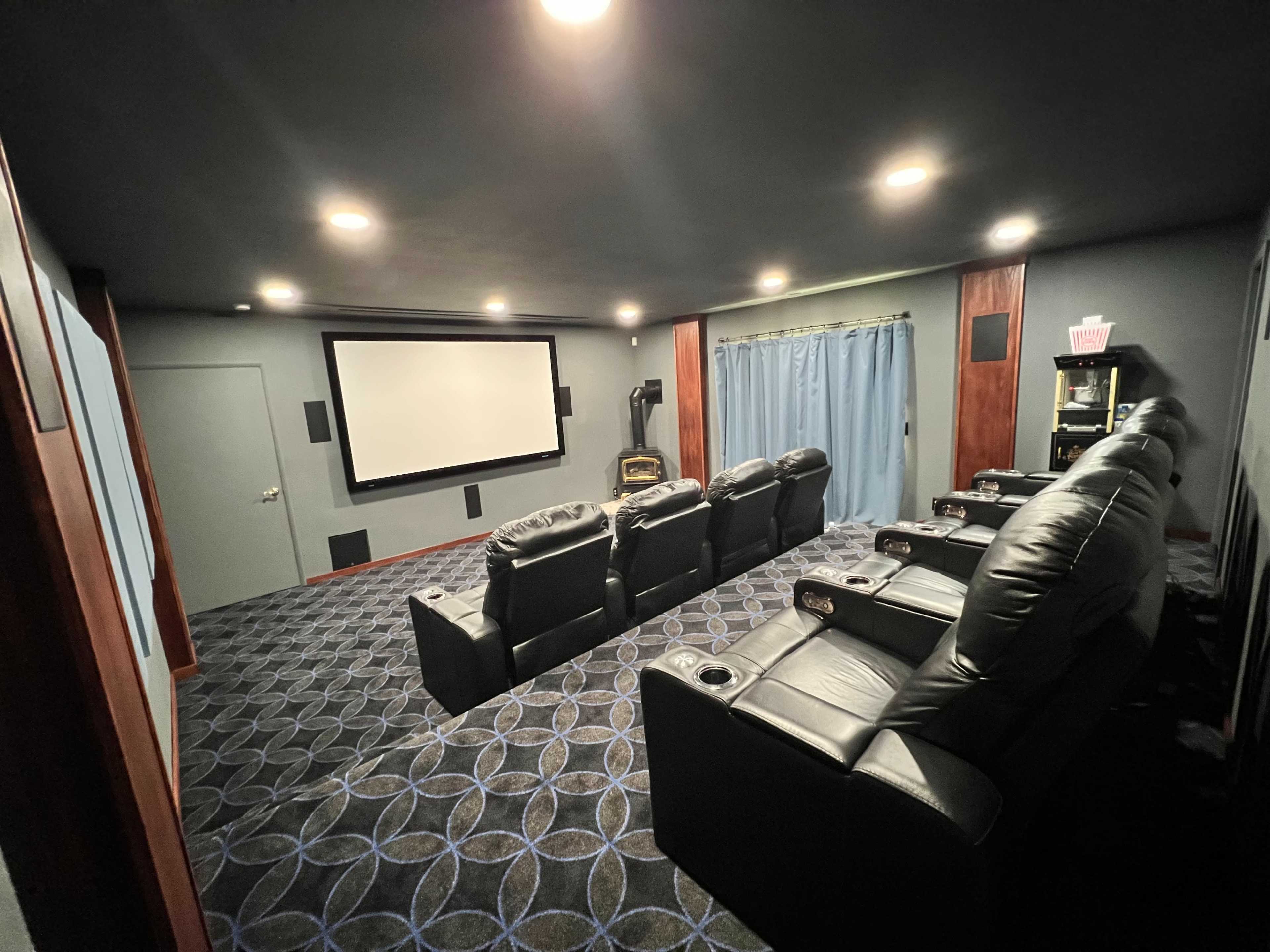 A home theater room featuring comfortable black recliner chairs arranged in front of a large projection screen, with blue curtains and a patterned carpet.