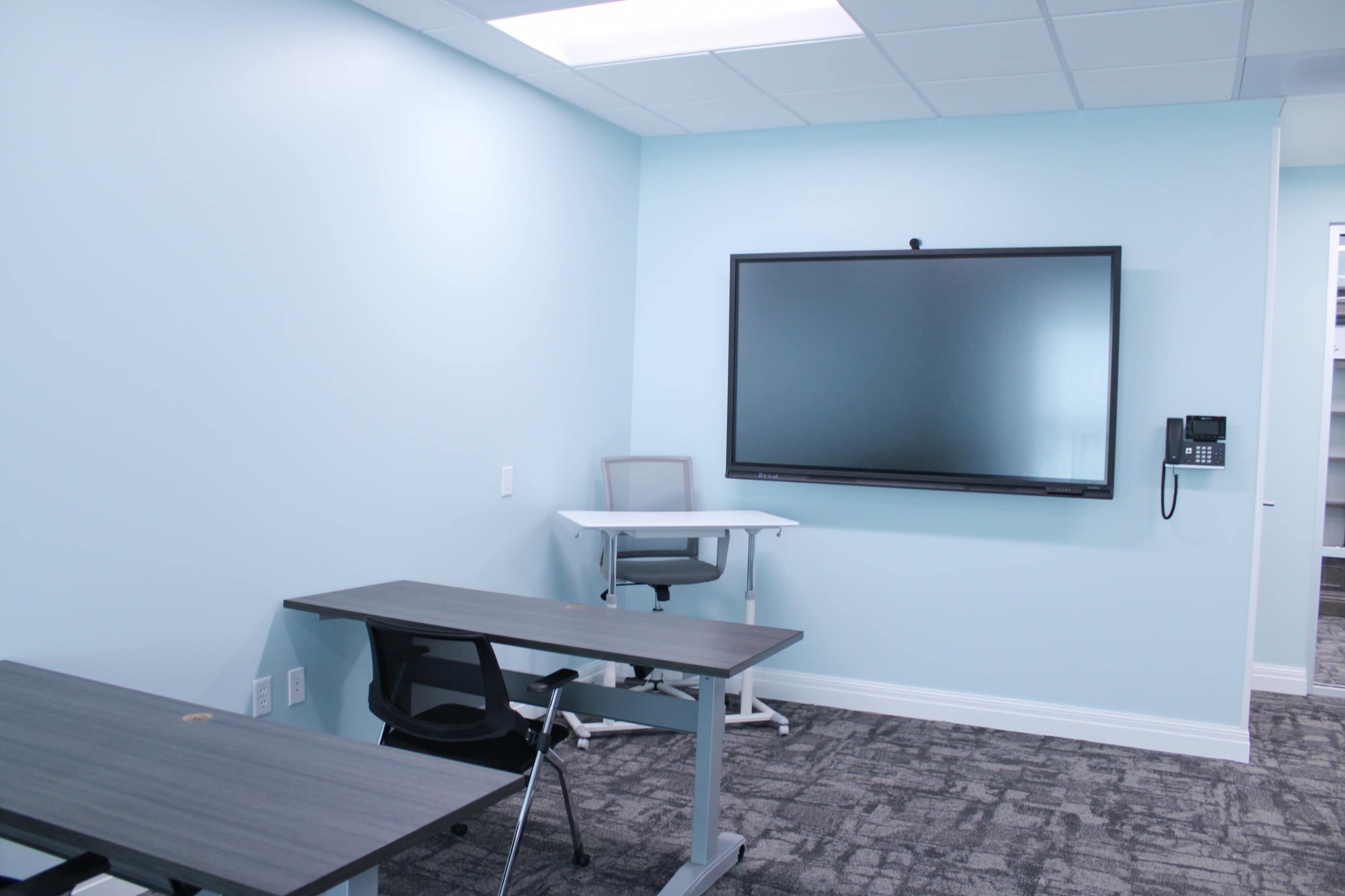 A small, brightly colored conference room with a large screen mounted on the wall, a desk, and a chair.
