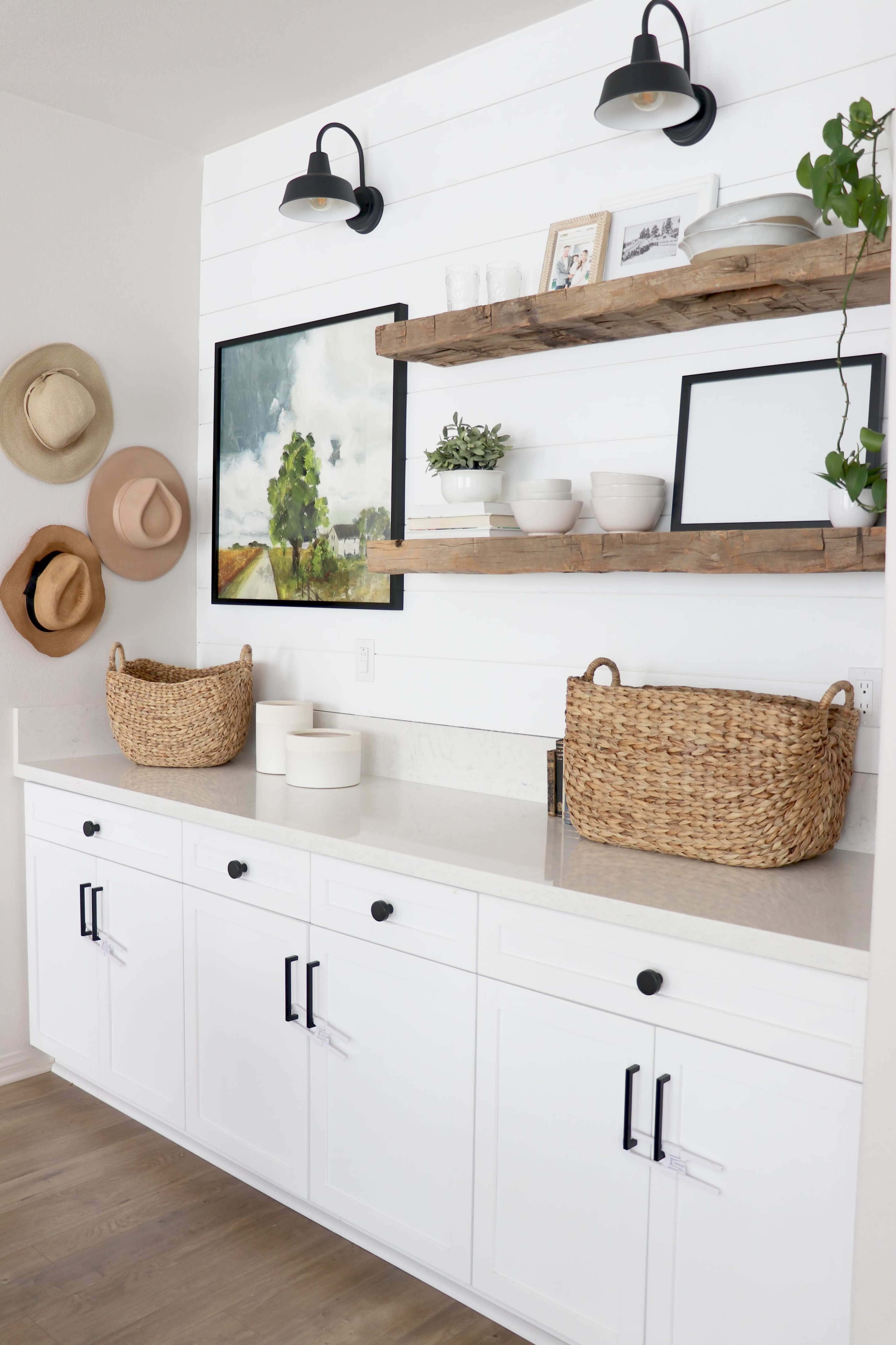 A stylish kitchen area featuring a white countertop with cabinets below, rustic shelves above displaying decorative items, and wall-mounted lights, alongside several hats hung on the adjacent wall.
