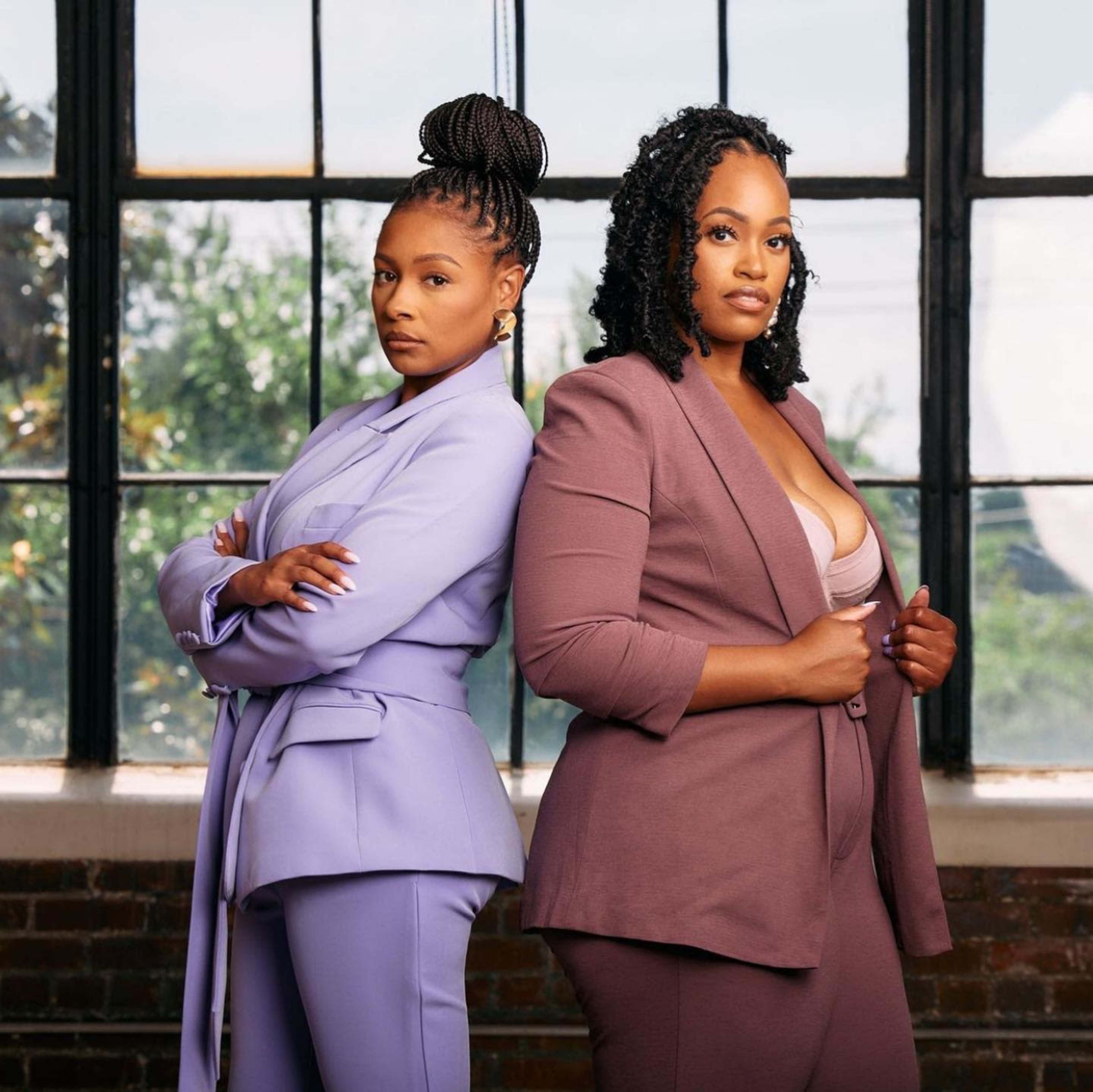 Two women in tailored suits stand back-to-back in front of large windows with greenery visible outside.