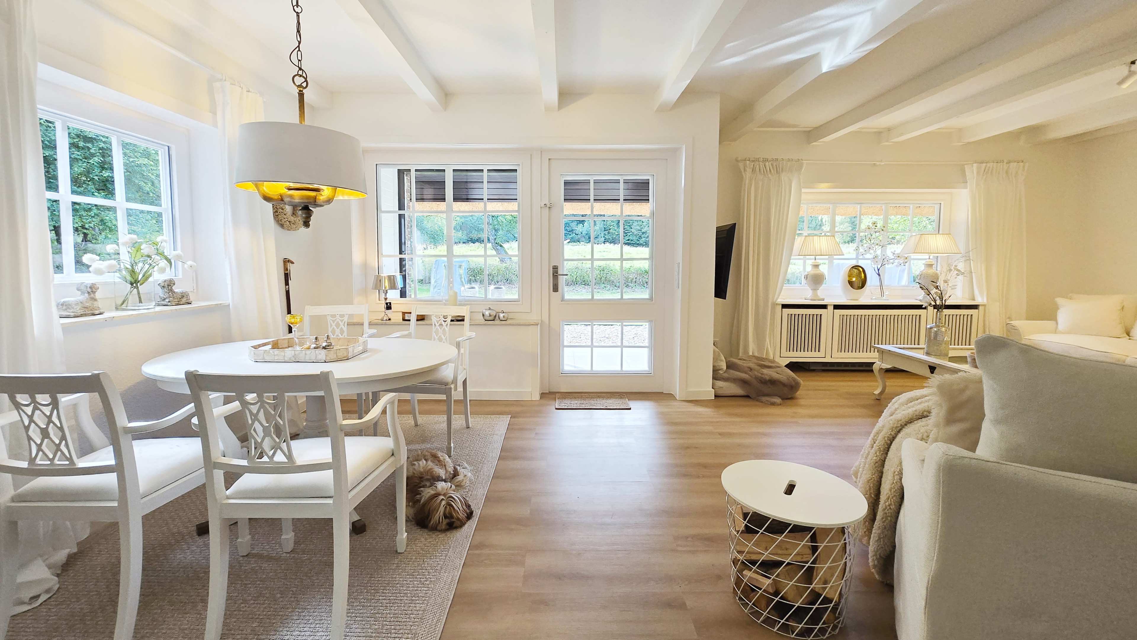 The image shows a bright, open living space featuring a dining area with a round table and four chairs, a cozy seating area, and large windows overlooking a garden.