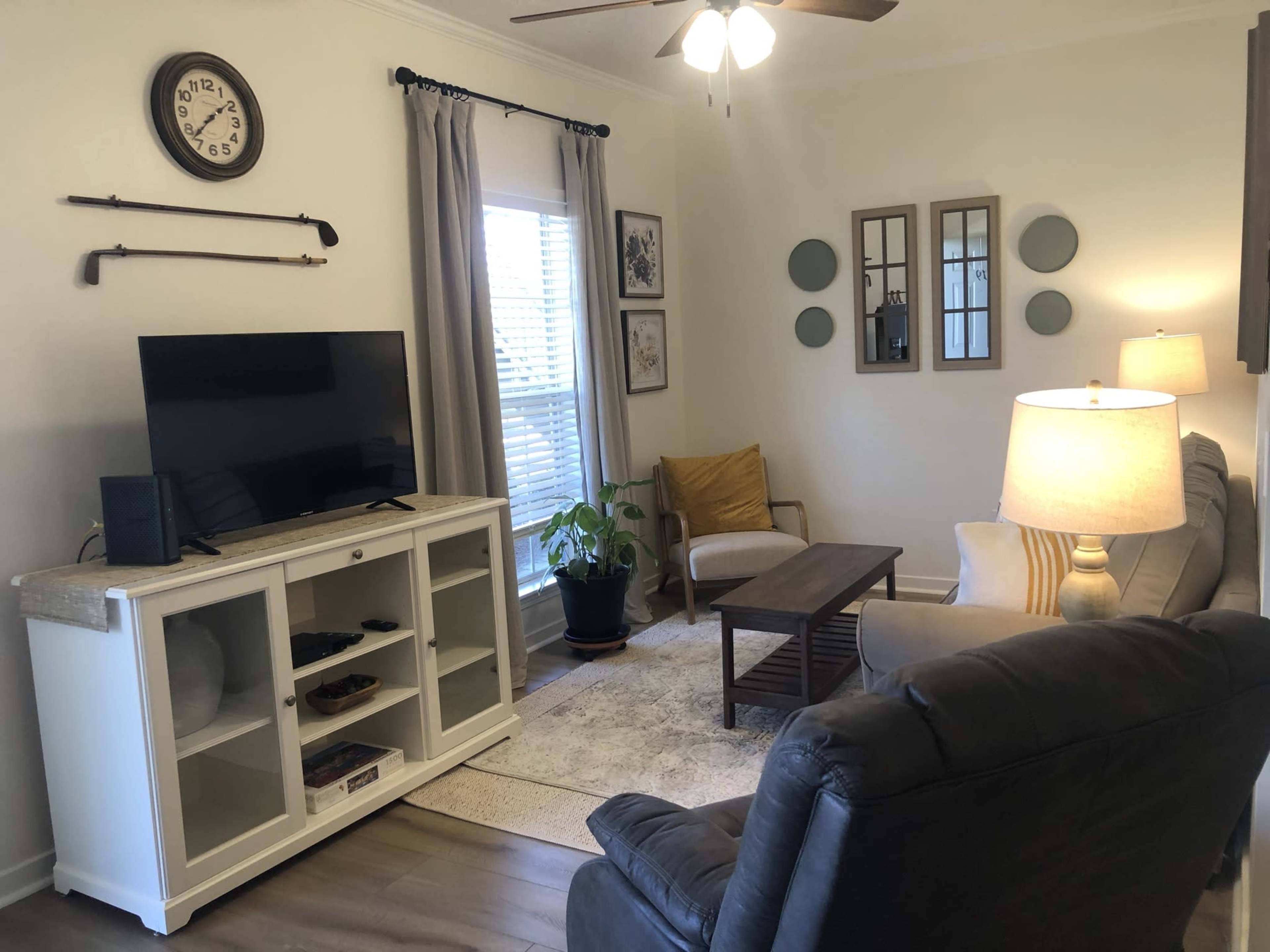 The living room features a television on a white cabinet, a coffee table, a light-colored sofa, and a comfortable chair, all arranged around a rug under a ceiling fan.