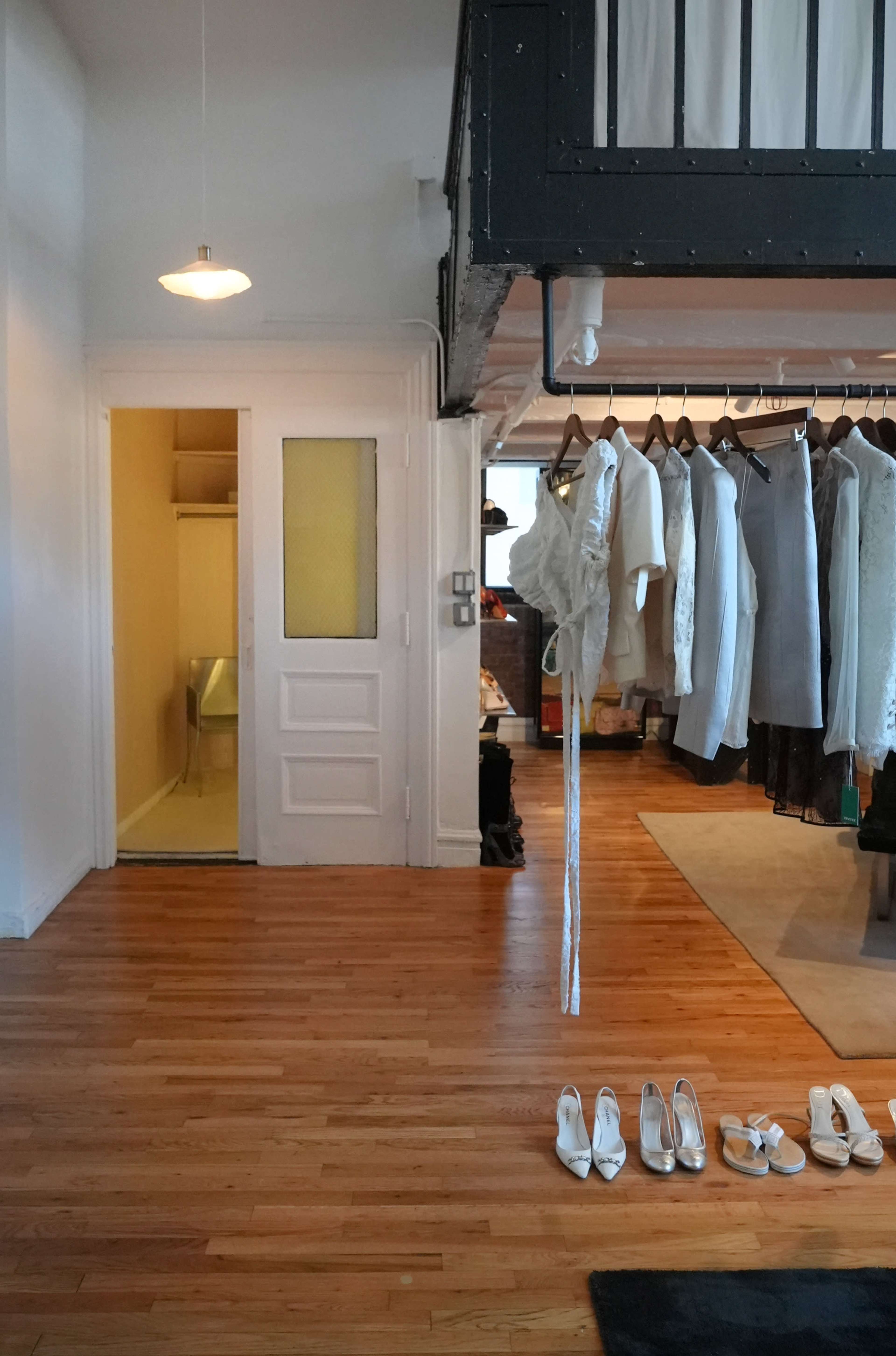 The image shows a wood-floored room with a white closet door on the left, a clothing rack filled with white garments, and a pair of white shoes neatly placed on the floor by a black mat.