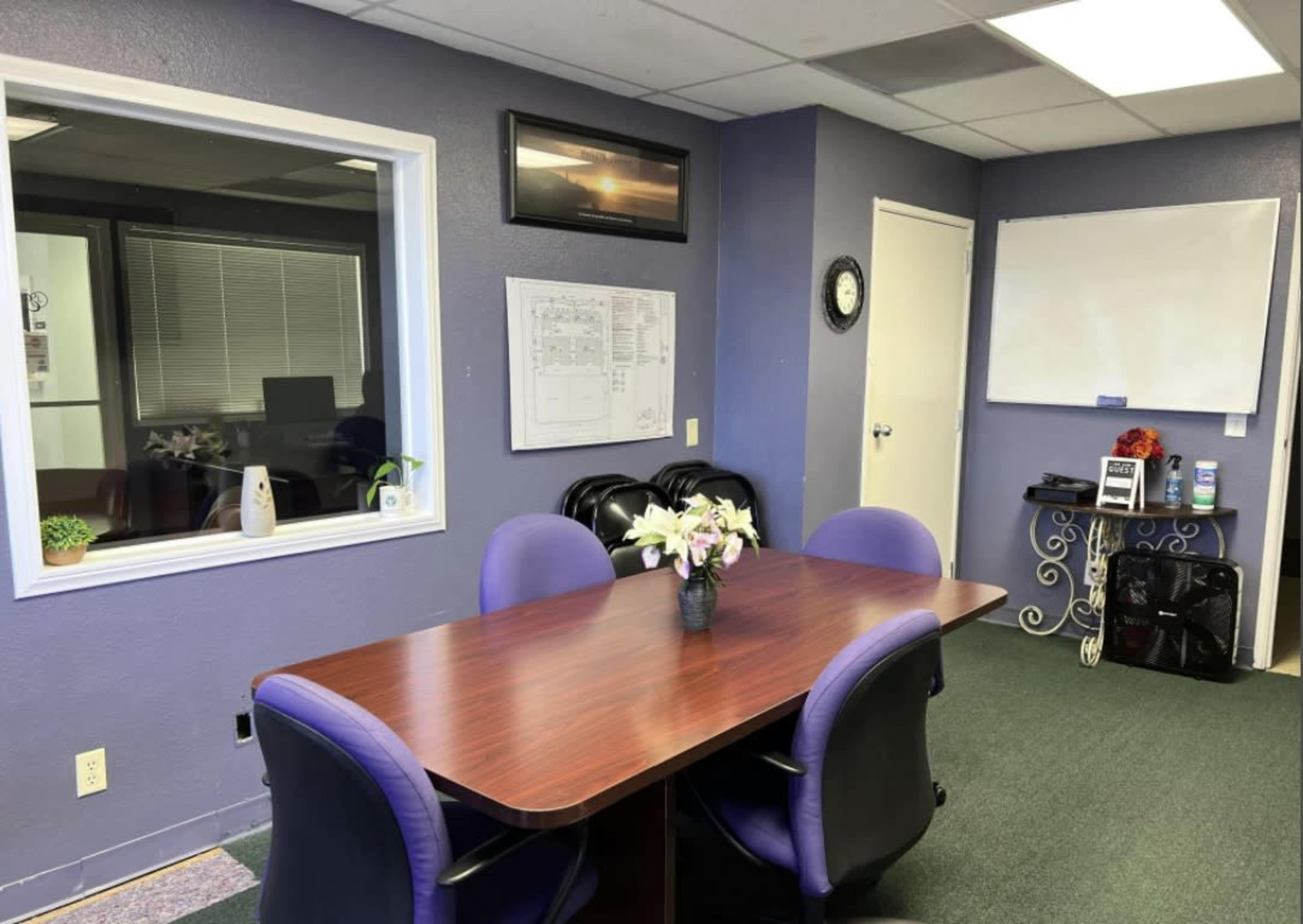 A meeting room features a brown table surrounded by black and purple chairs, with a window, a wall-mounted TV, a whiteboard, and various office supplies.