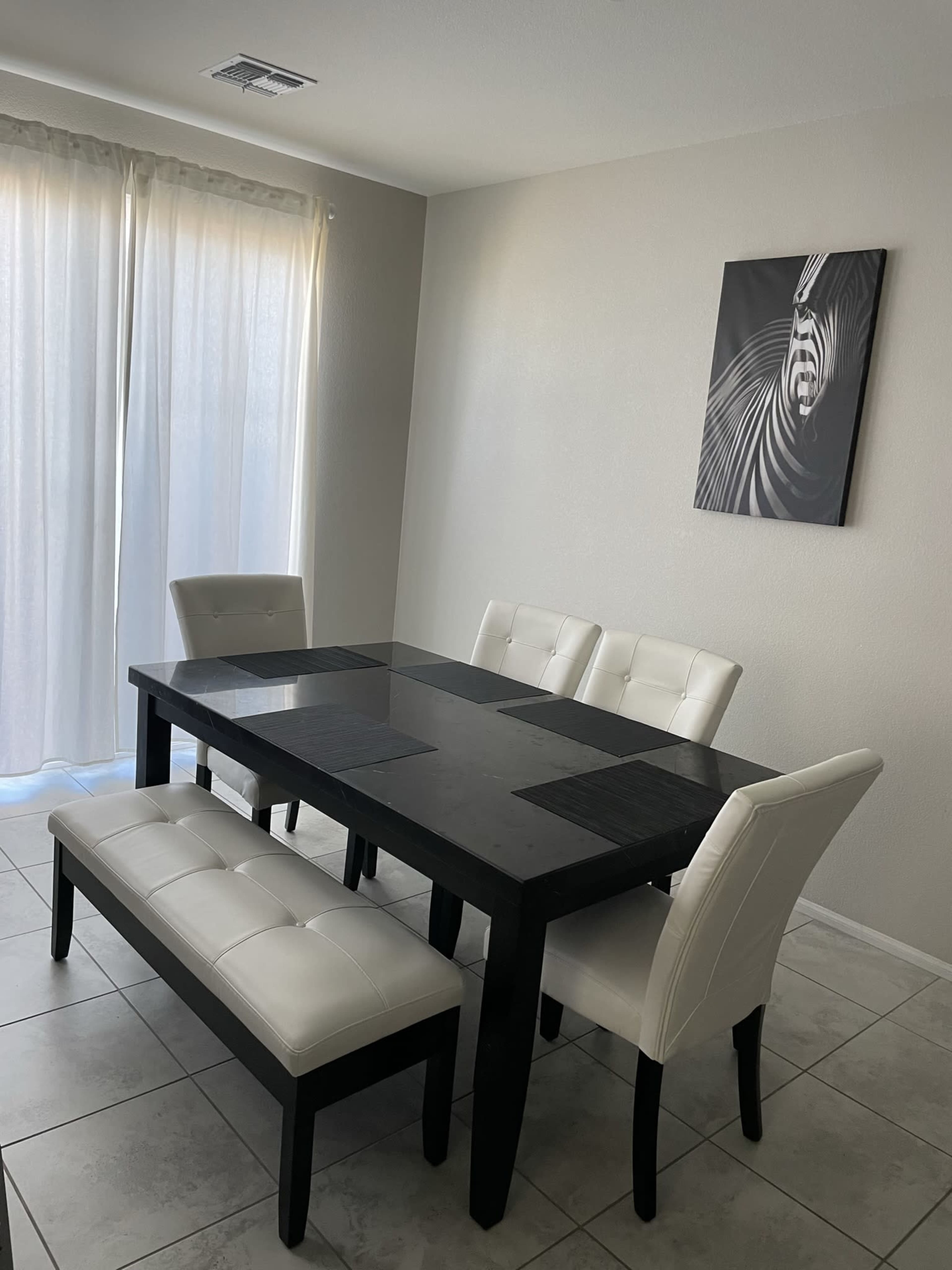 The image shows a dining area with a black table, four white chairs, and a bench, illuminated by natural light from a nearby window.