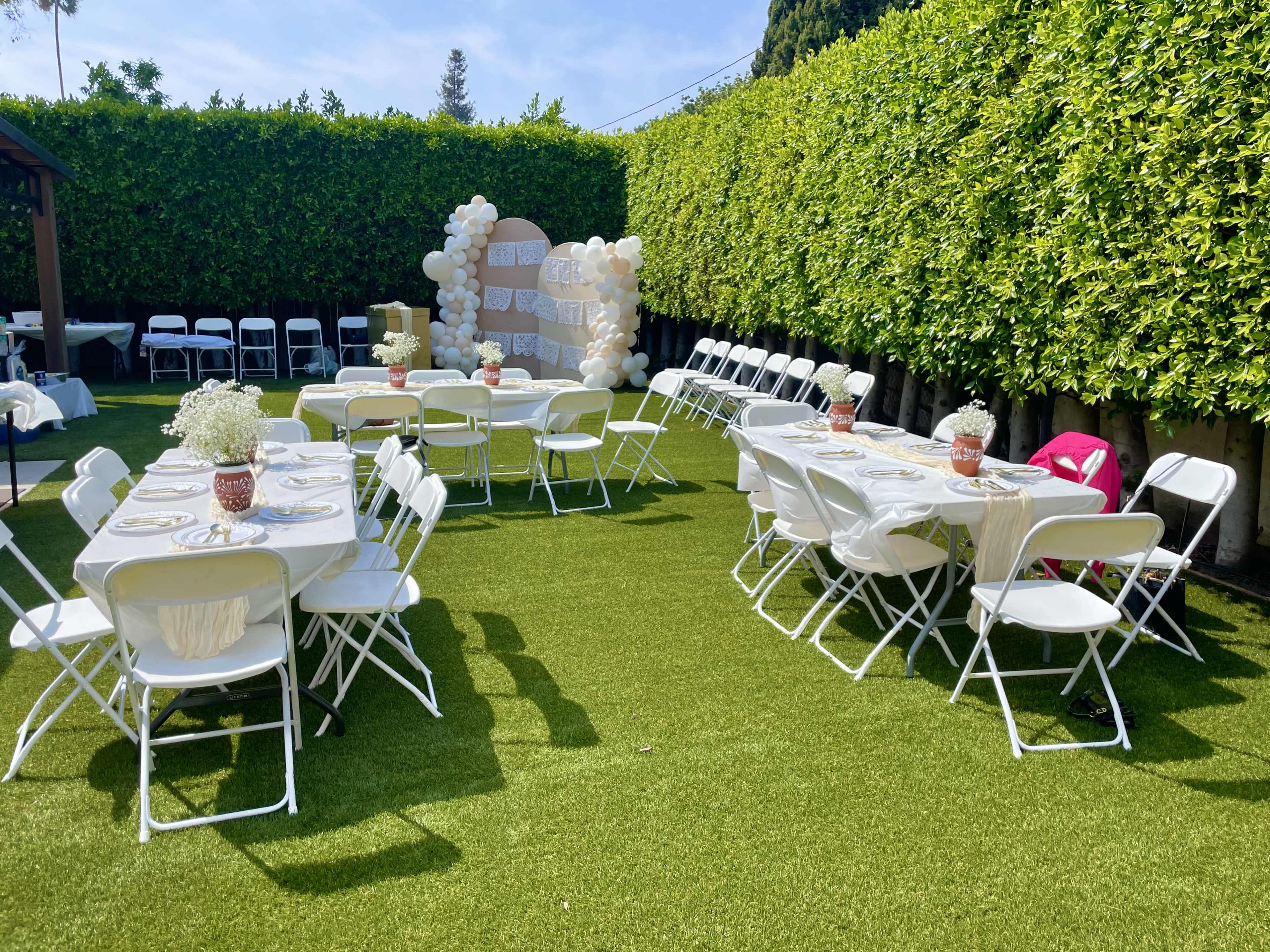 The image shows a neatly arranged outdoor event space with white tables and chairs set up on green turf, surrounded by tall hedges and decorated with balloons.