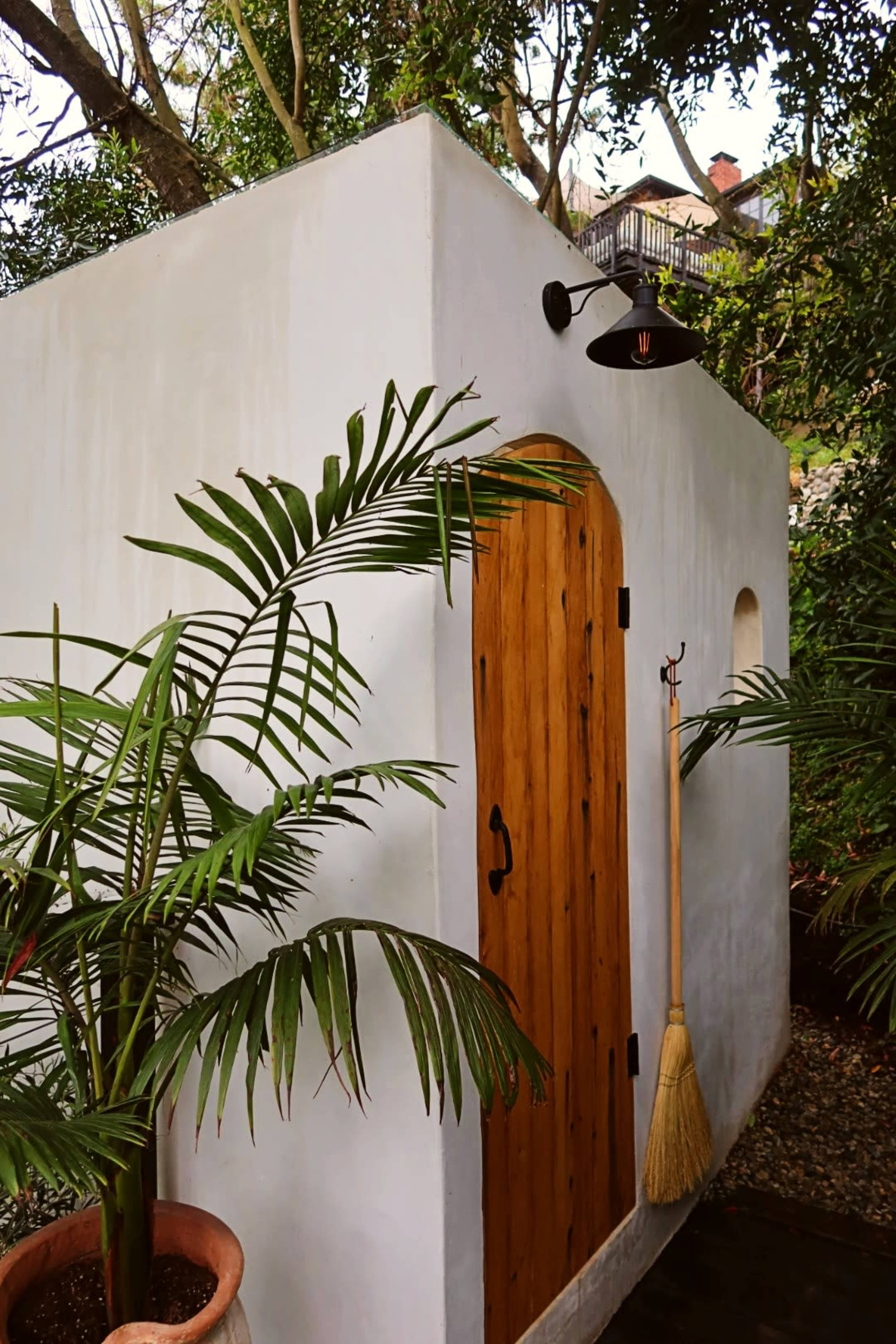 A white-walled structure with a wooden door is accented by a wall-mounted light and surrounded by tropical plants.
