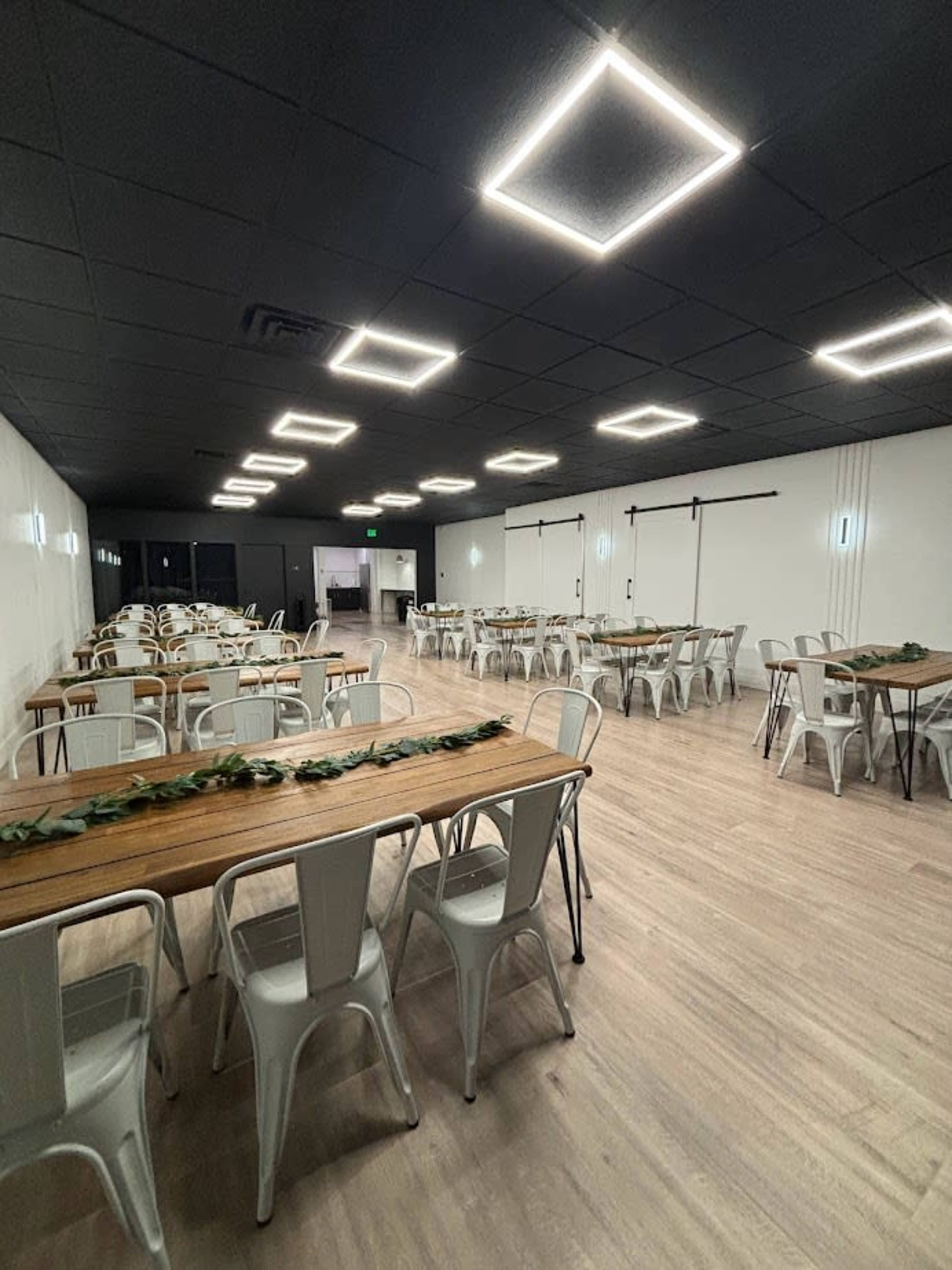 A spacious event room featuring several long wooden tables surrounded by metal chairs, illuminated by square ceiling lights.