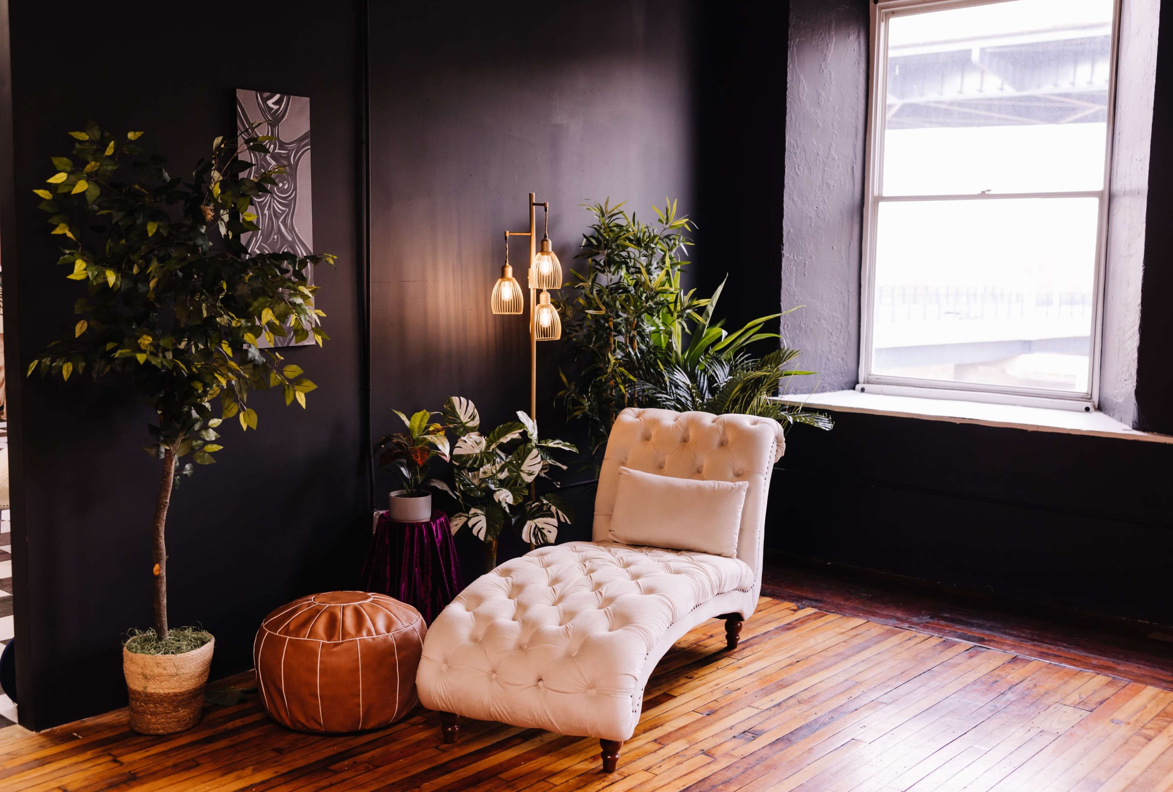 The image shows a stylish interior featuring a tufted cream chaise lounge, a brown pouf, and various green plants beside a large window.