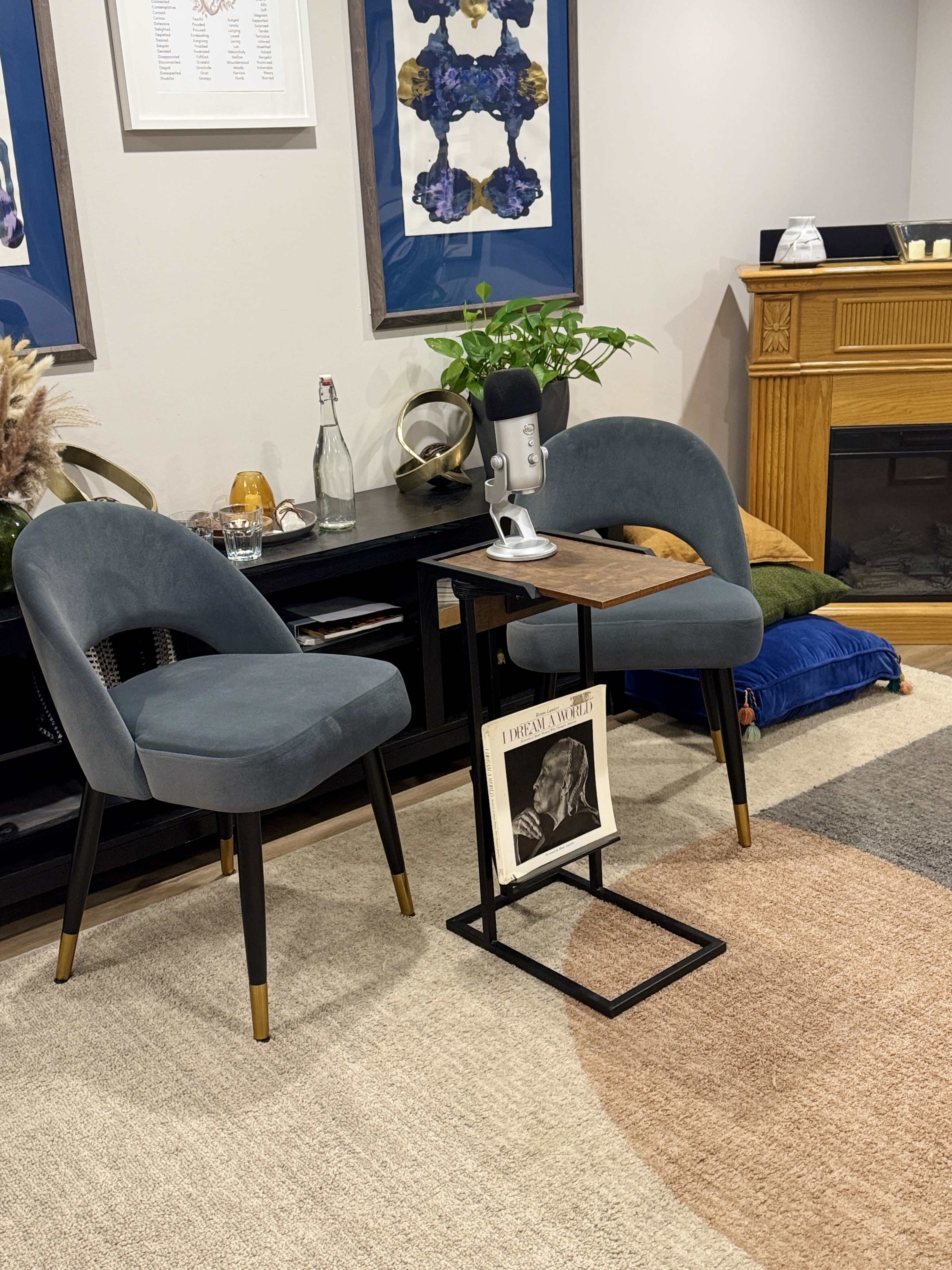 The image shows a cozy interior setting with two gray chairs, a small wooden table holding a microphone, and a framed magazine on the floor, all arranged near a decorative fireplace.