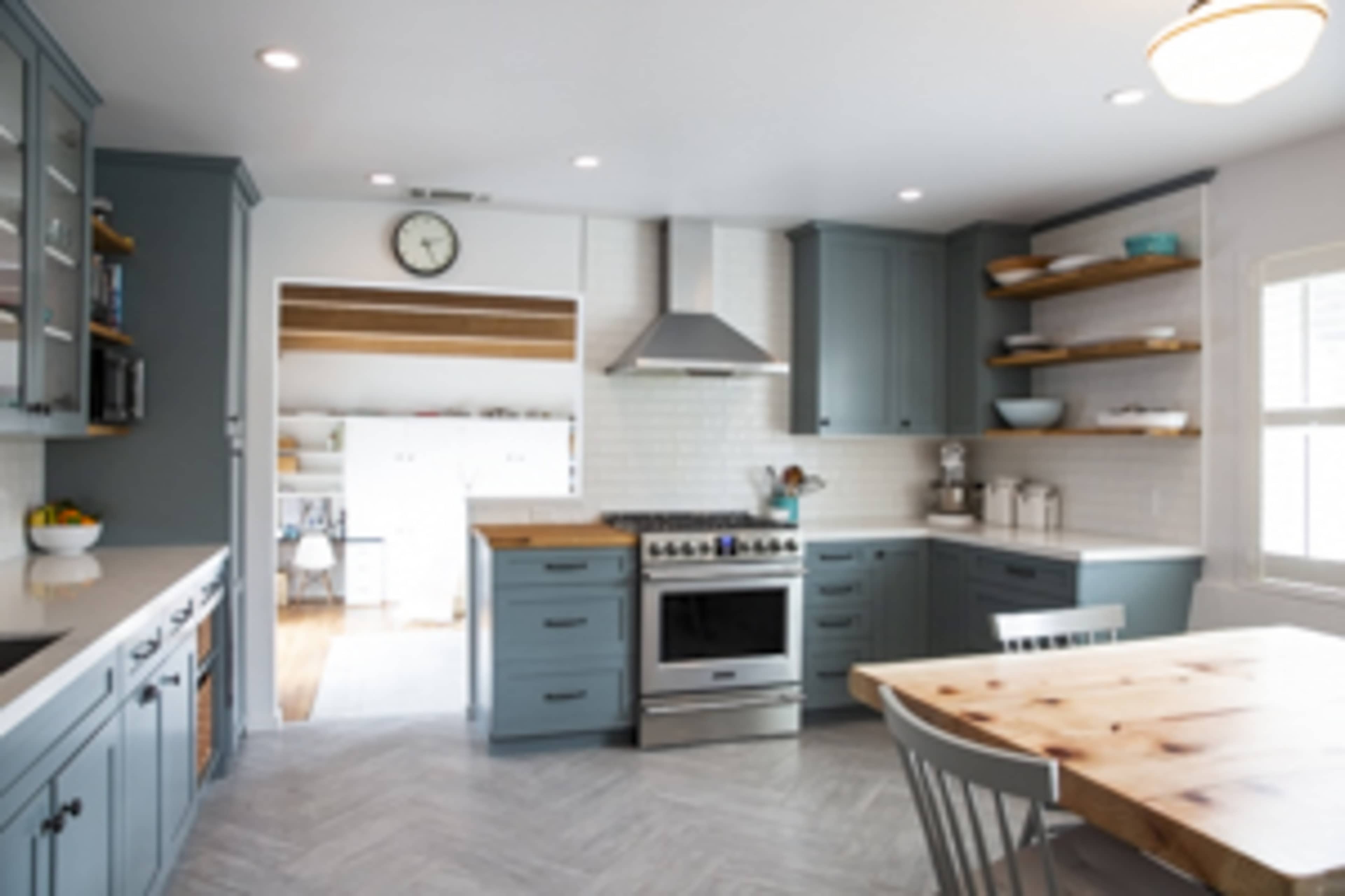A modern kitchen features sleek blue cabinets, a stainless steel oven, and a wooden dining table with white chairs.