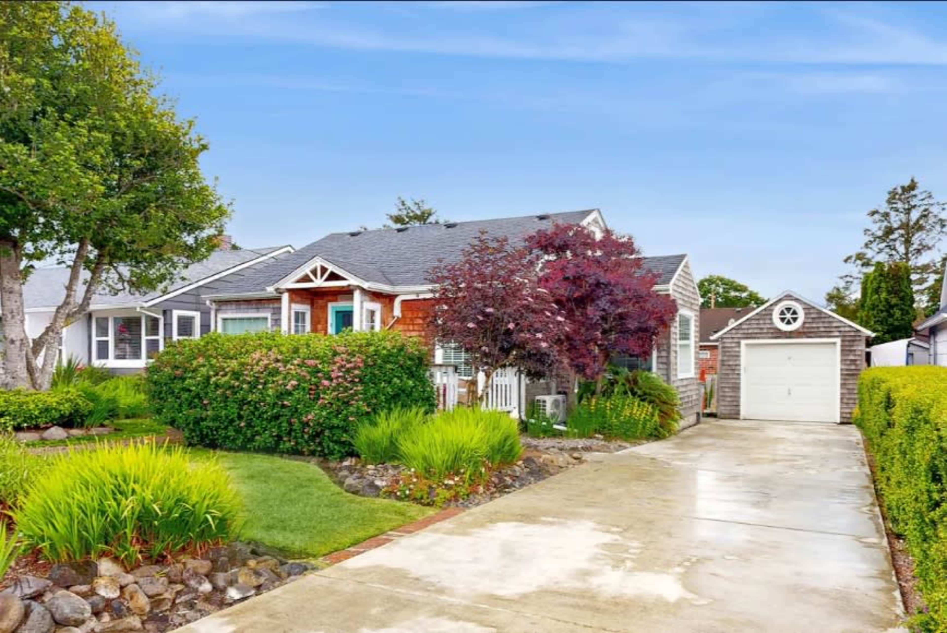 A single-story house with a well-maintained lawn and garden, accompanied by a garage on the side, is set against a clear blue sky.