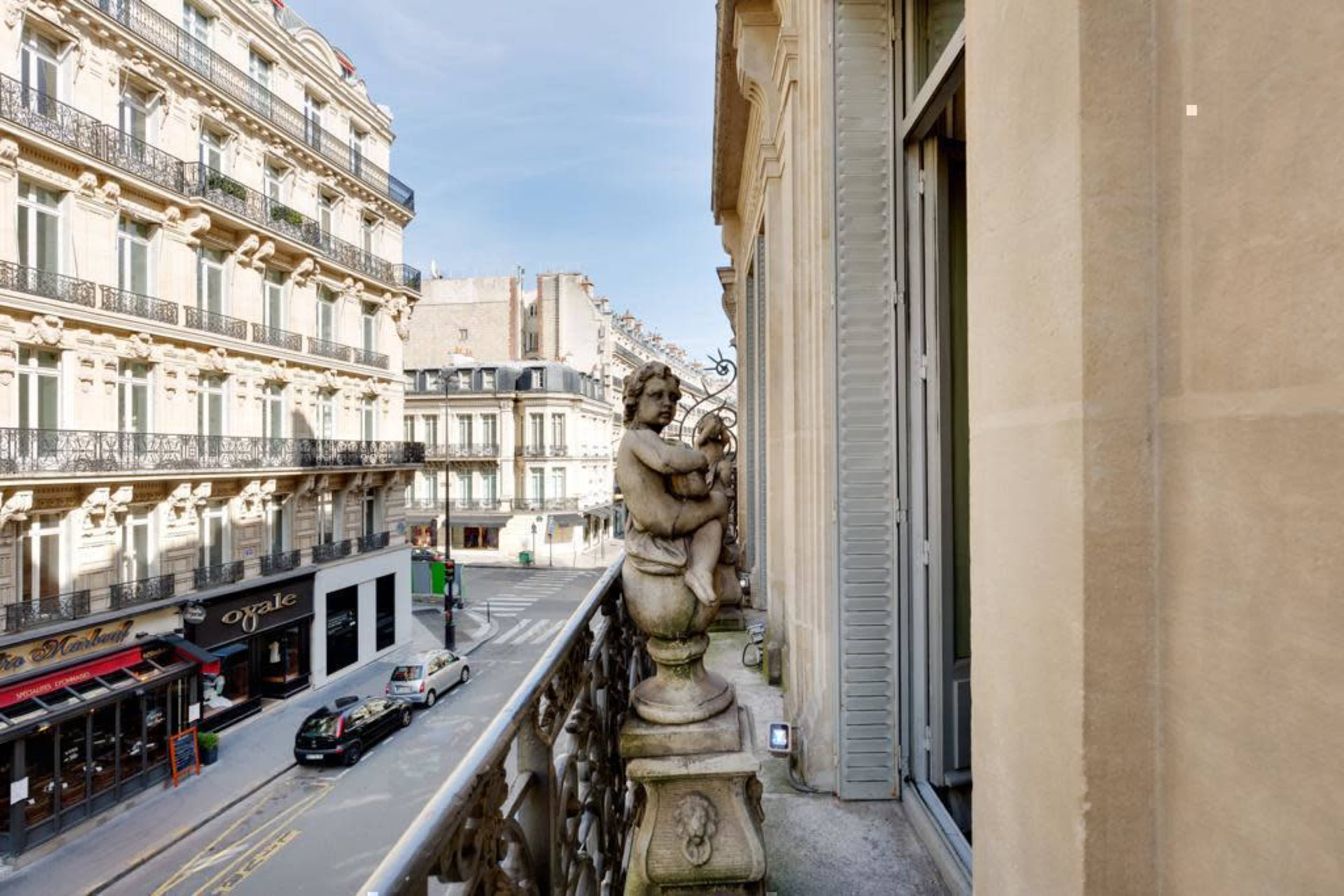 A stone statue of a cherub stands on a balcony overlooking a street lined with buildings in Paris.