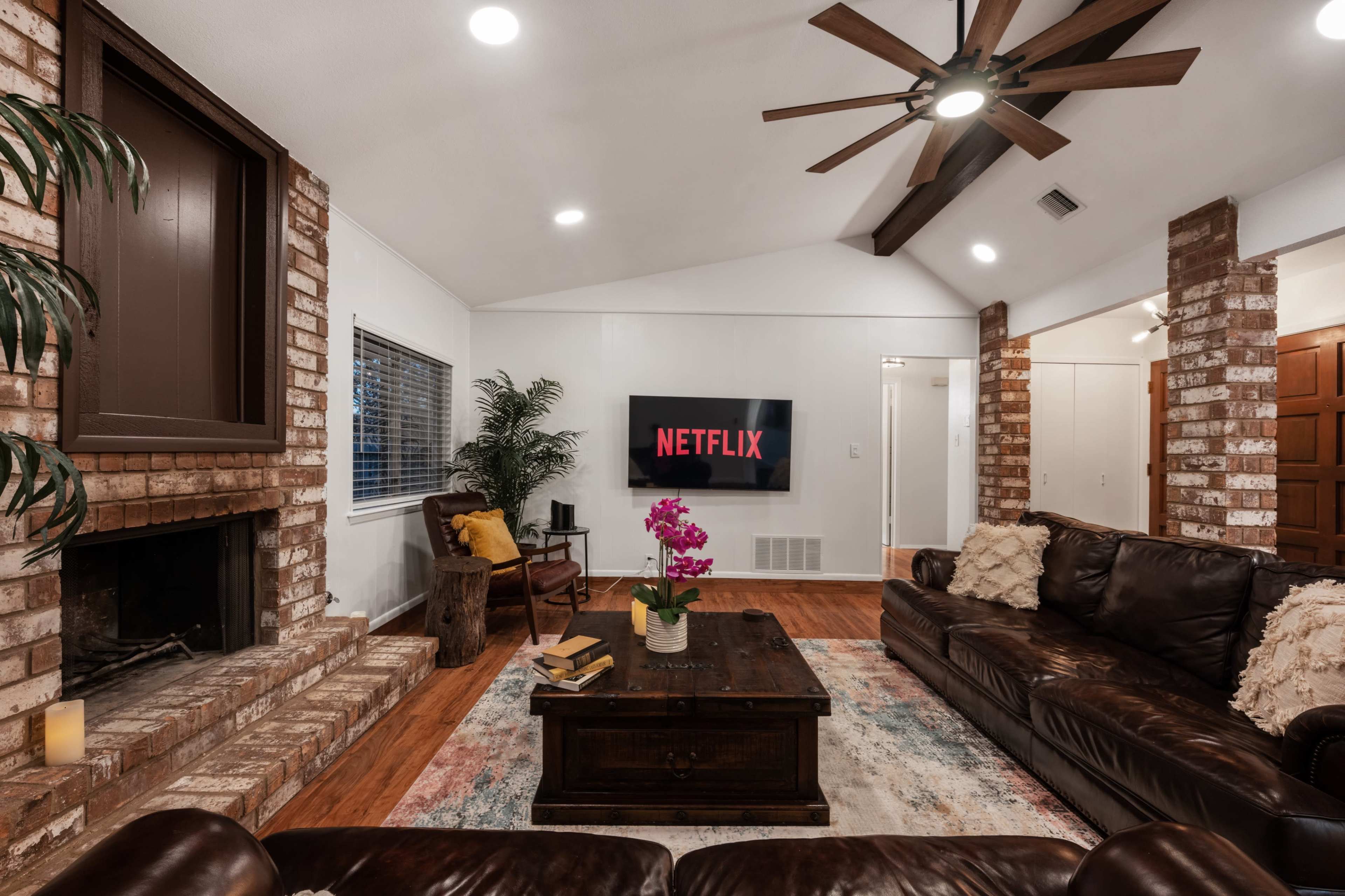A cozy living room features a brick fireplace, a large wooden coffee table, a Netflix display on the wall, and leather sofas.