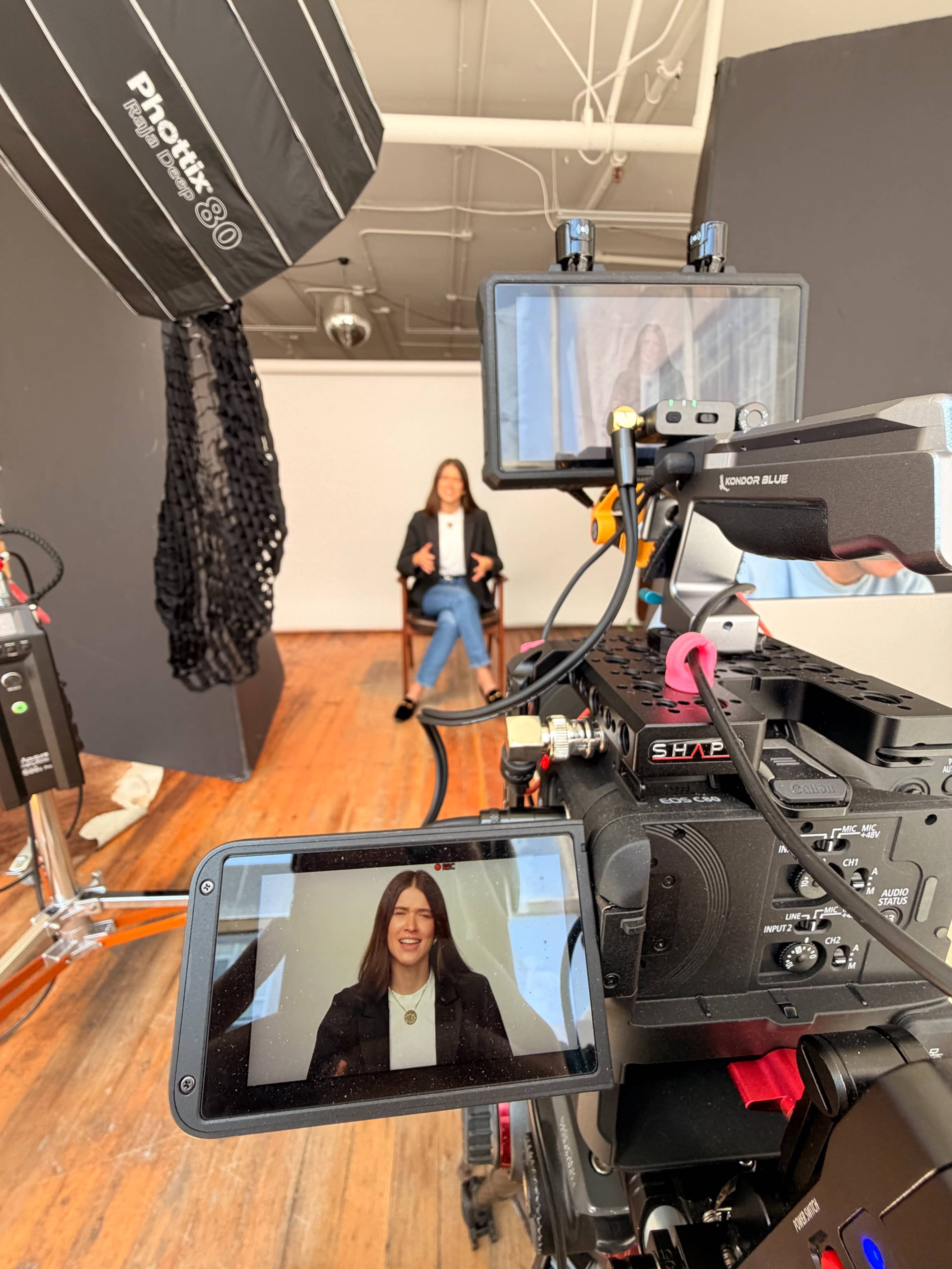 The image shows a woman seated in a director’s chair in a studio, with camera equipment and lighting set up in the foreground.