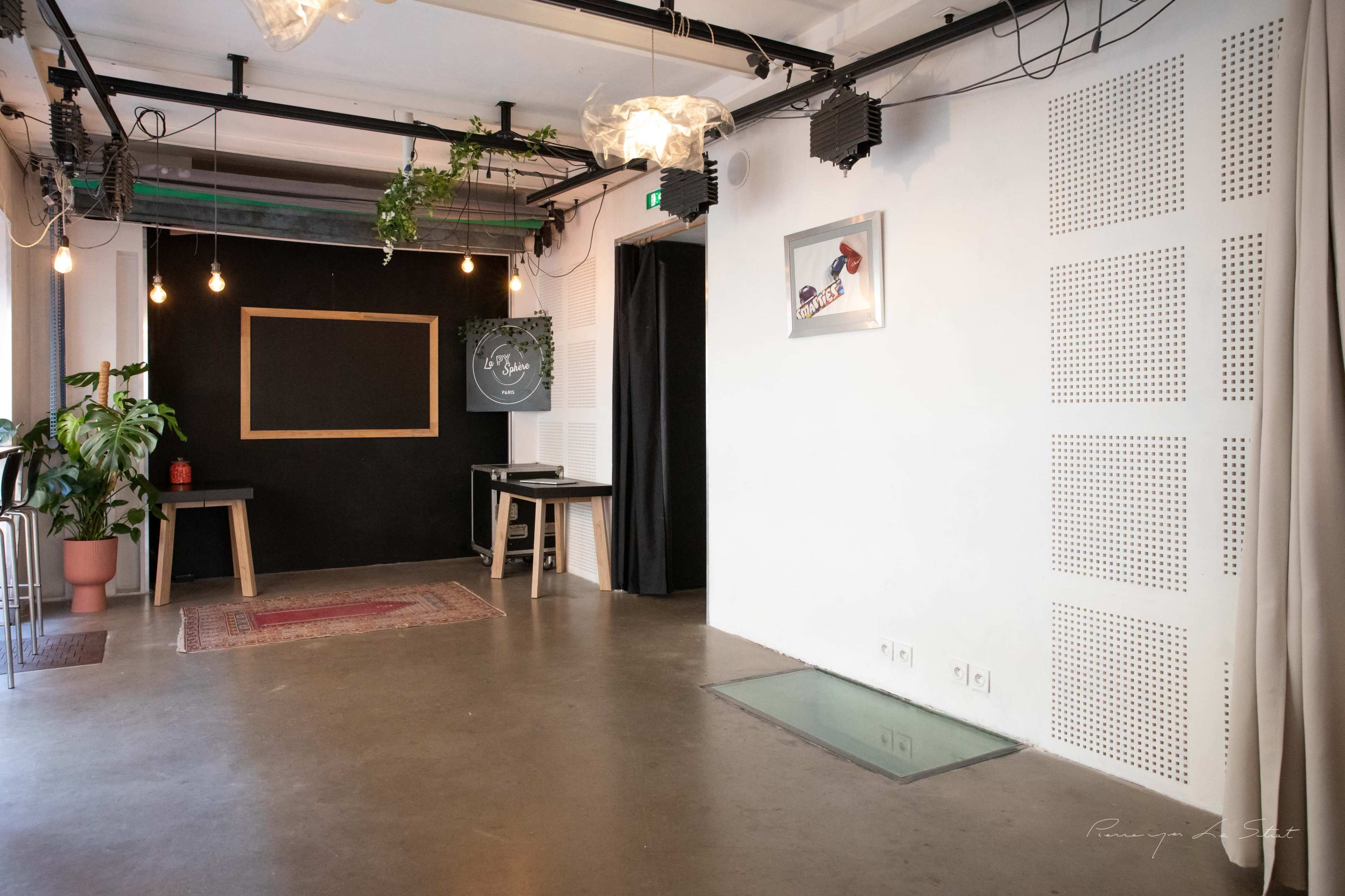 The image depicts an empty, modern interior space featuring a black wall with a chalkboard, a potted plant, and exposed light bulbs hanging from the ceiling.