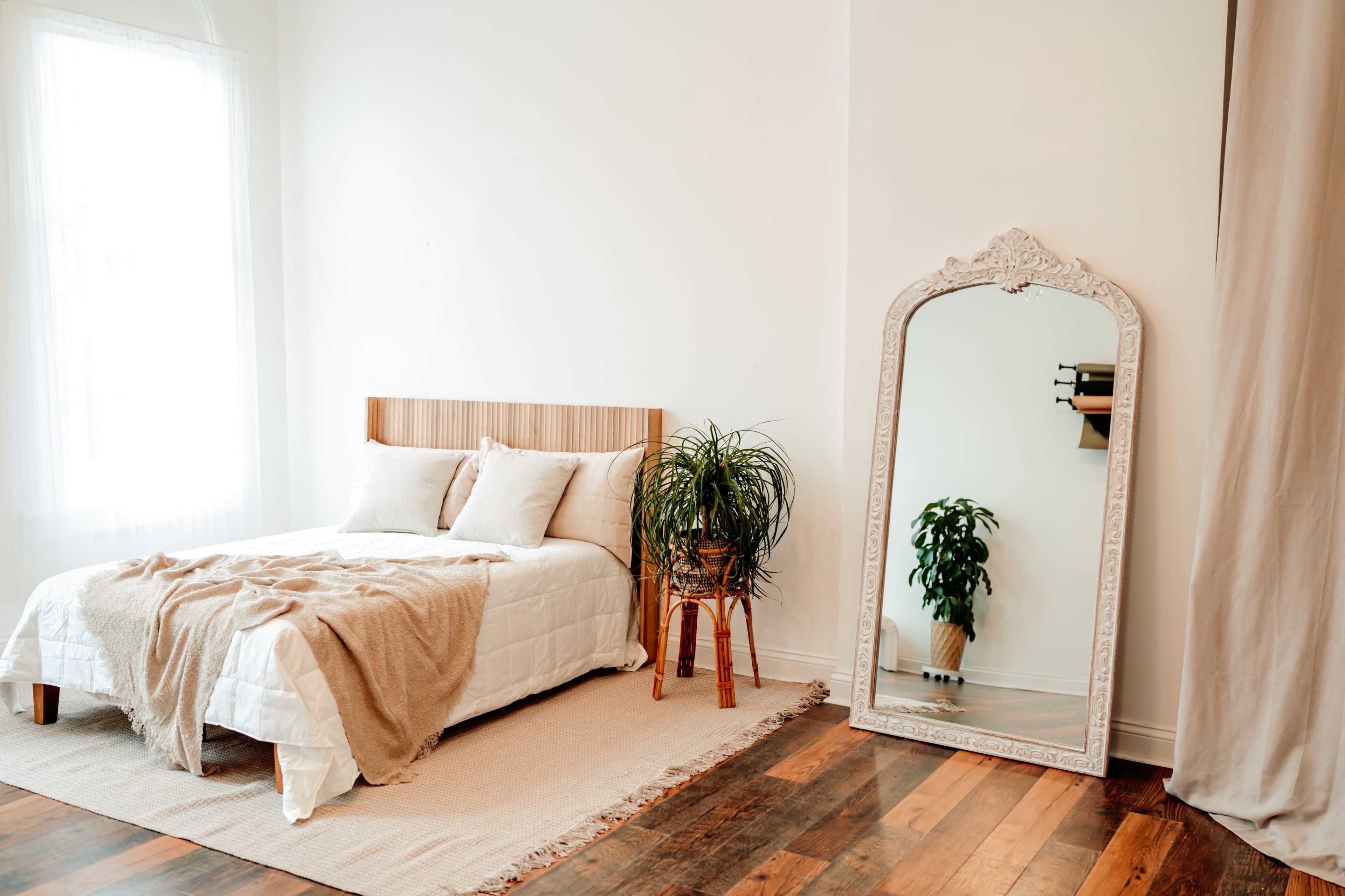 A simple bedroom with a wooden bed, a large ornate mirror, a potted plant, and a rug on wooden flooring.