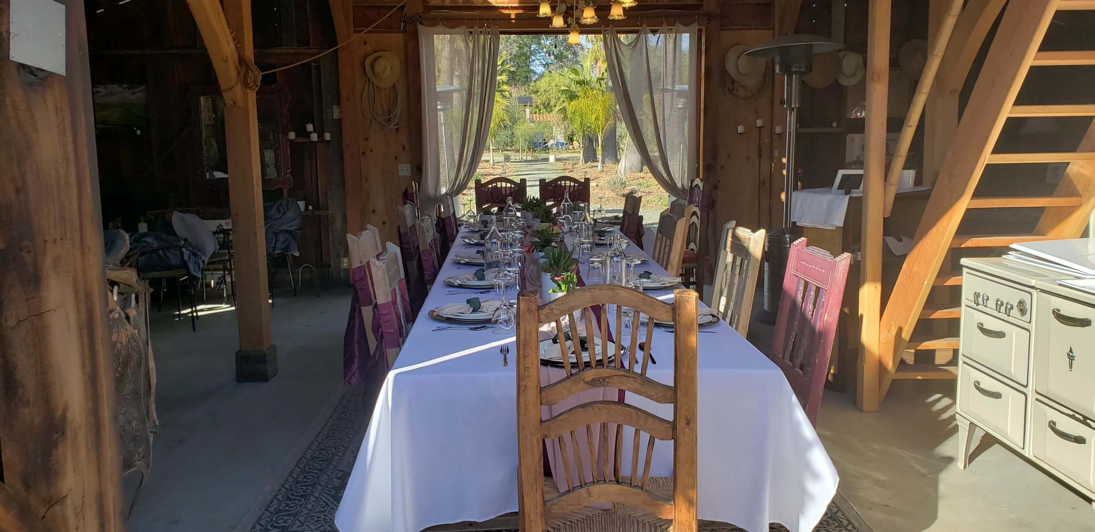A long dining table is set with plates and decorations inside a rustic barn, with wooden chairs along the sides and a view of greenery through large windows.