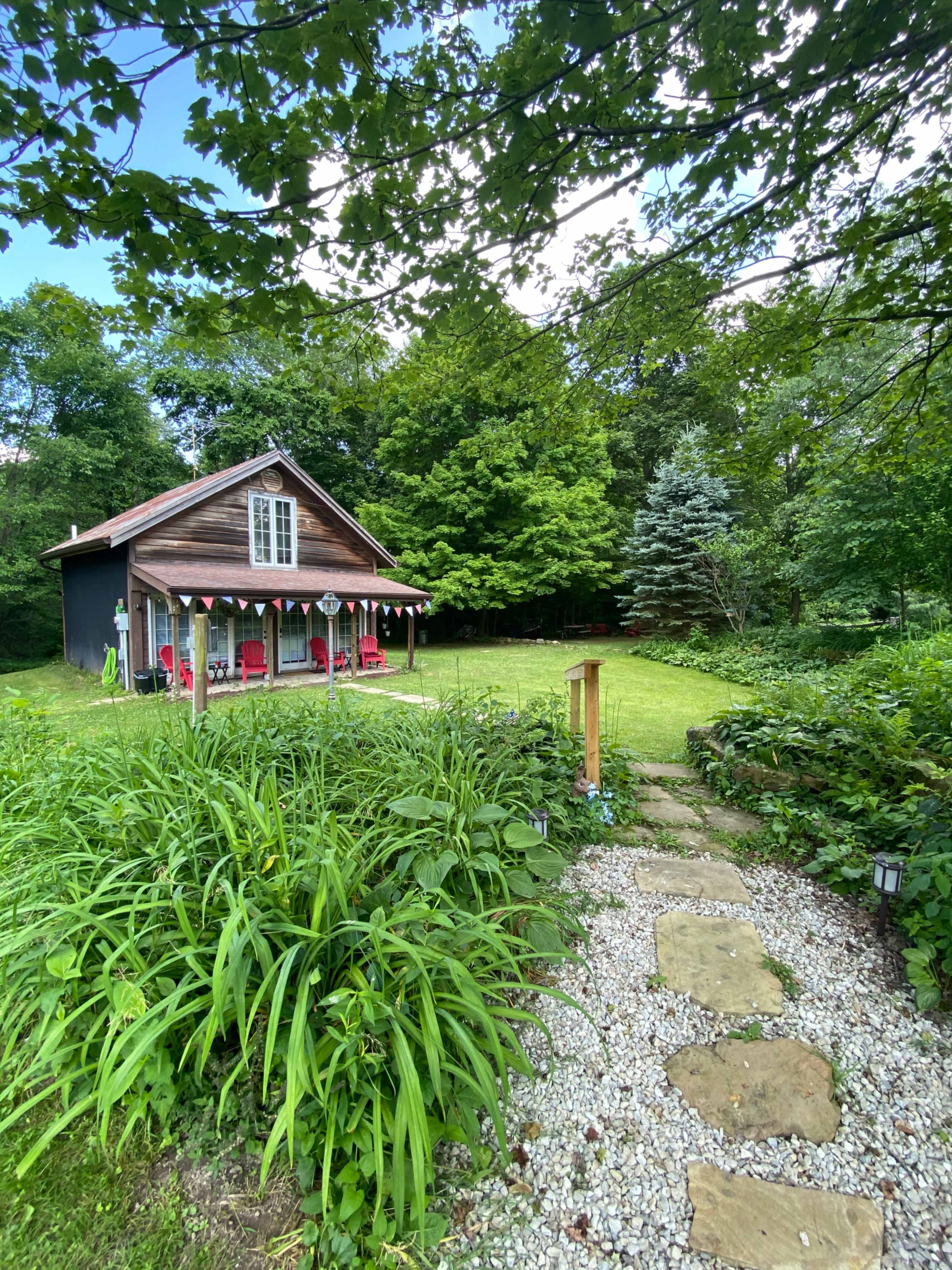 A stone pathway leads through lush greenery to a wooden house with a porch decorated with chairs and bunting.
