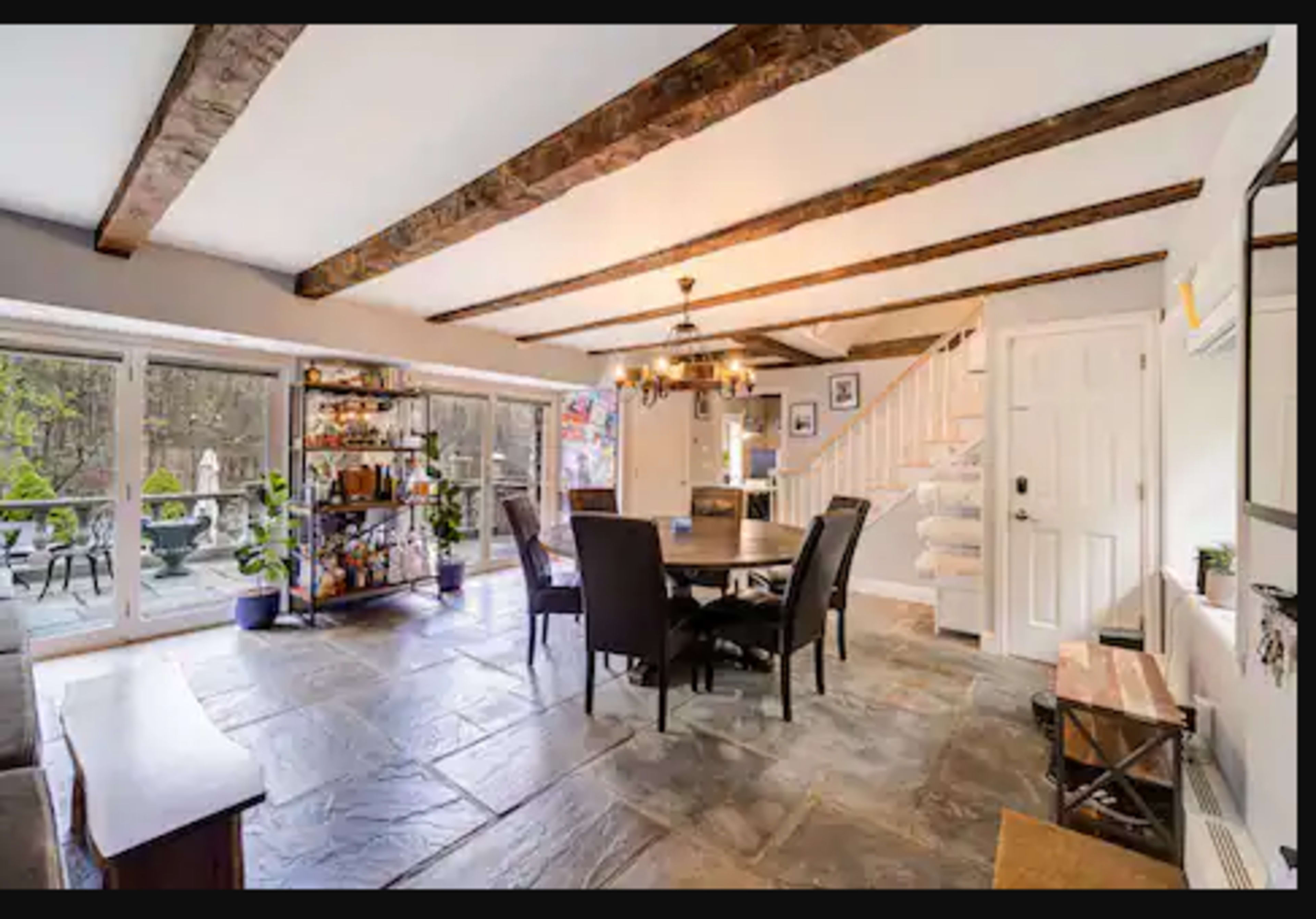 A cozy dining area with a round table, black chairs, and a staircase, all set against a backdrop of large windows and exposed wooden beams.