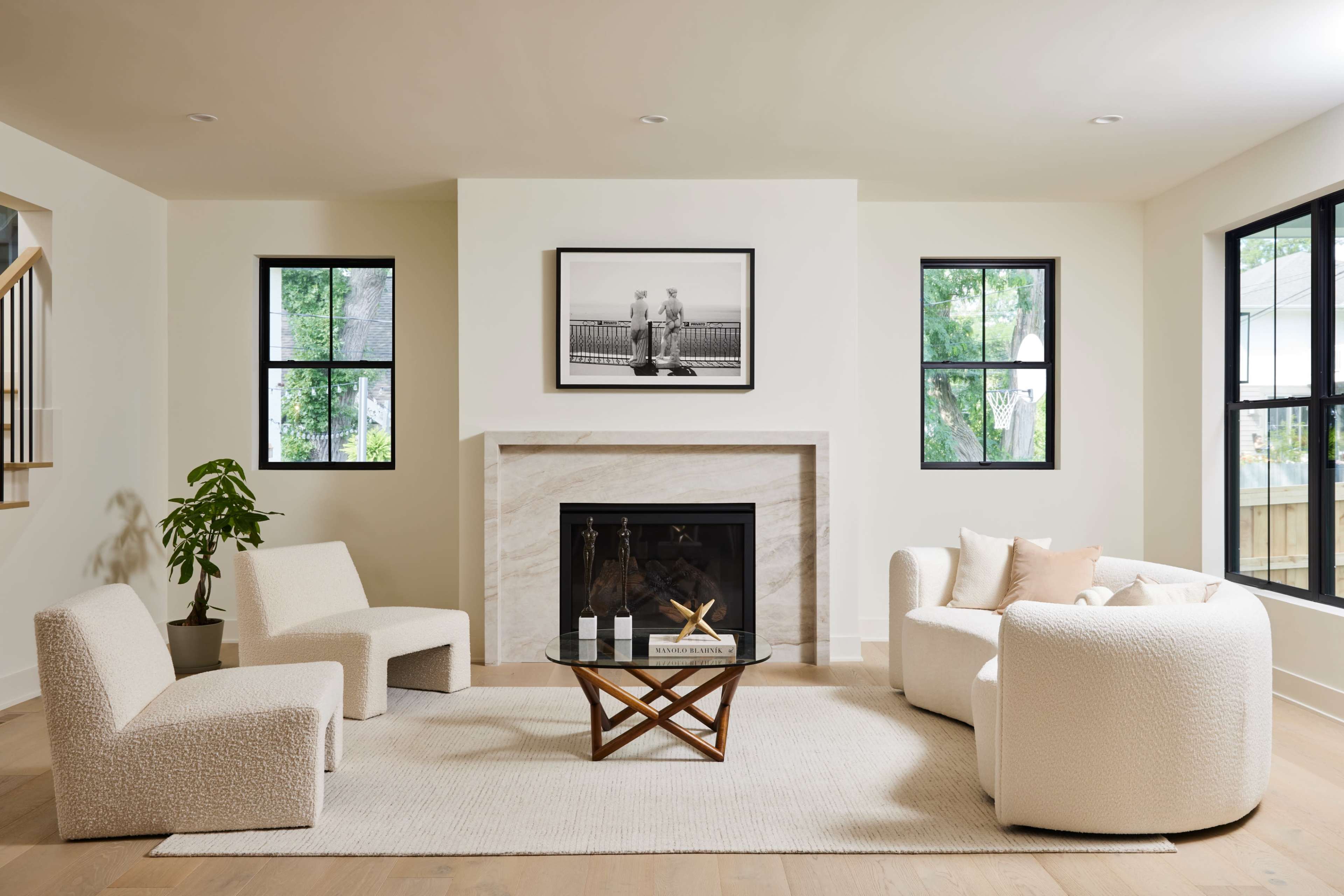 A modern living room features two upholstered chairs, a round sofa, a glass coffee table on a rug, and a fireplace with a framed black-and-white photograph above it.