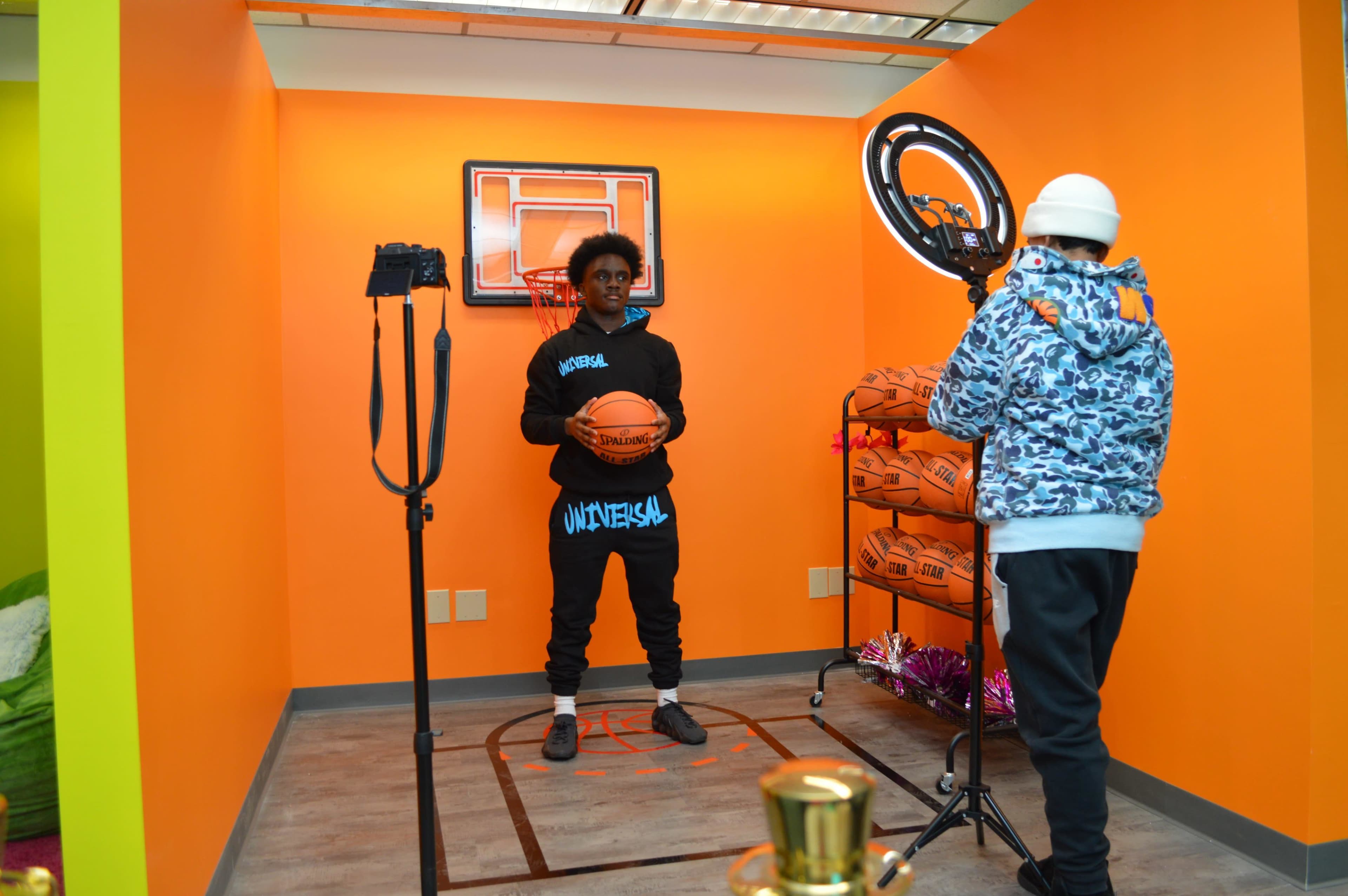 A person stands holding a basketball in front of a small basketball hoop set against an orange wall, while another person adjusts a camera on a tripod.