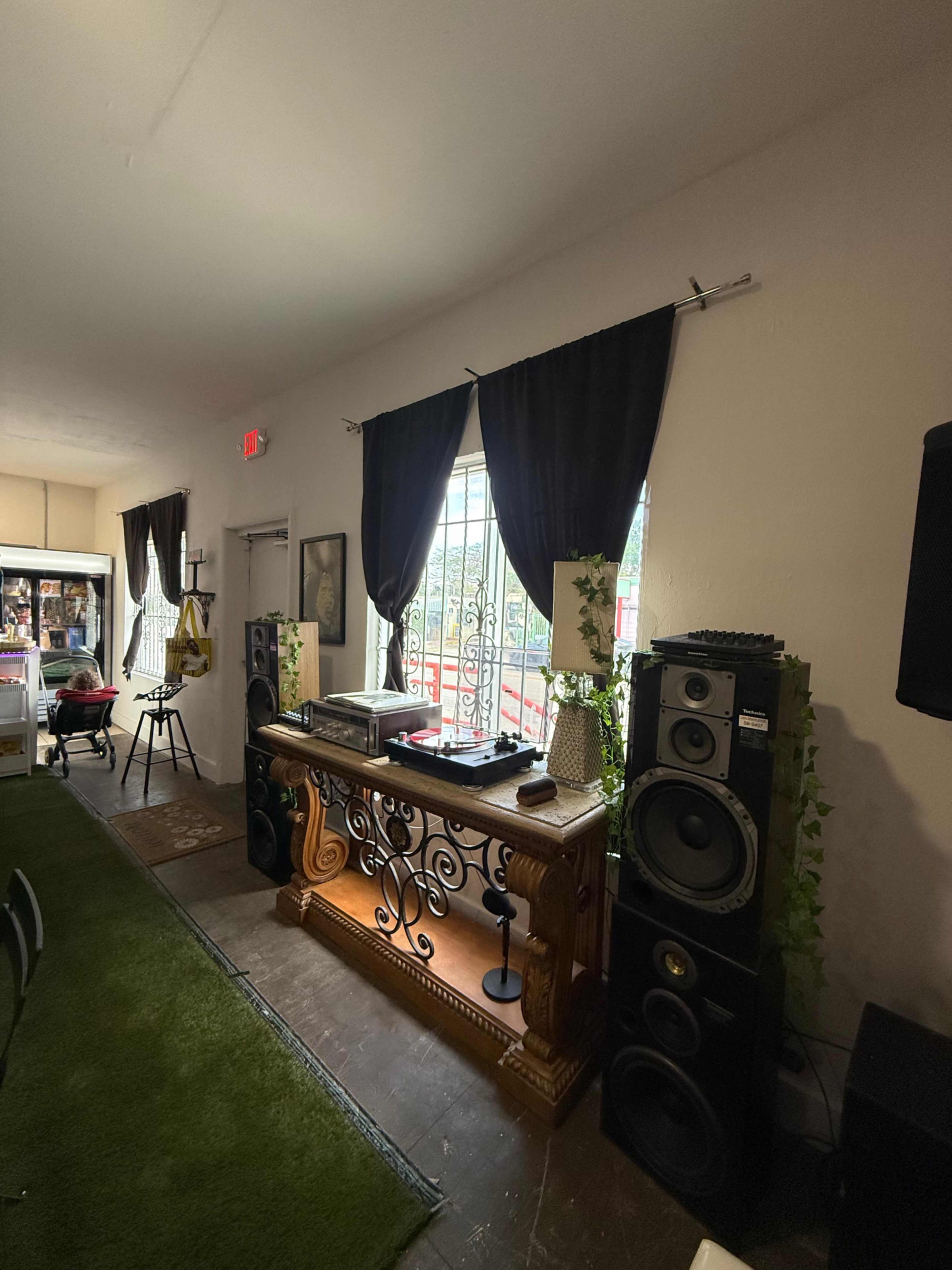 The image shows a room with a DJ setup, featuring a turntable on a decorative wooden table, flanked by large speaker systems against a wall with windows covered by dark curtains.