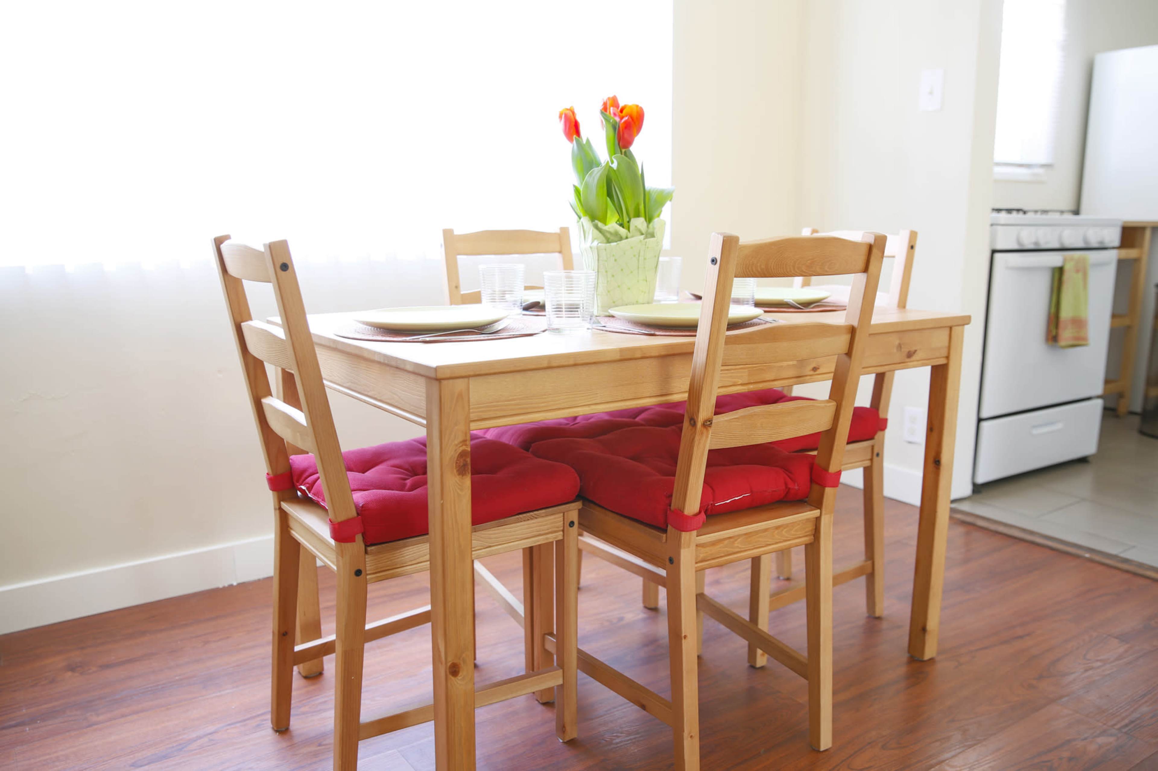 A wooden dining table with four chairs, each featuring red seat cushions, is set for a meal with plates and glasses, alongside a vase of tulips on the table.