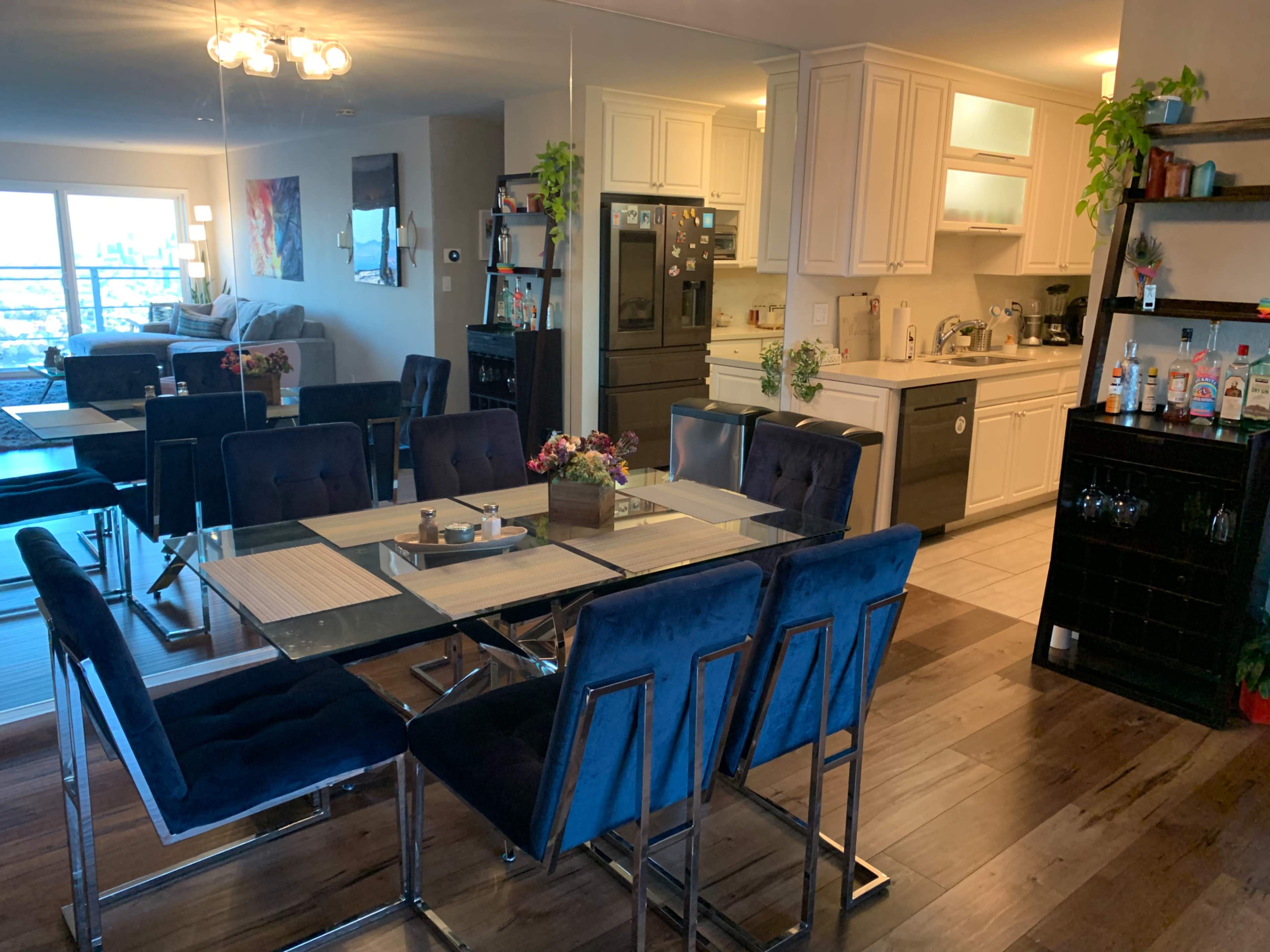 A dining area featuring a glass-top table surrounded by blue upholstered chairs, with an open kitchen in the background and a living area visible through a large window.
