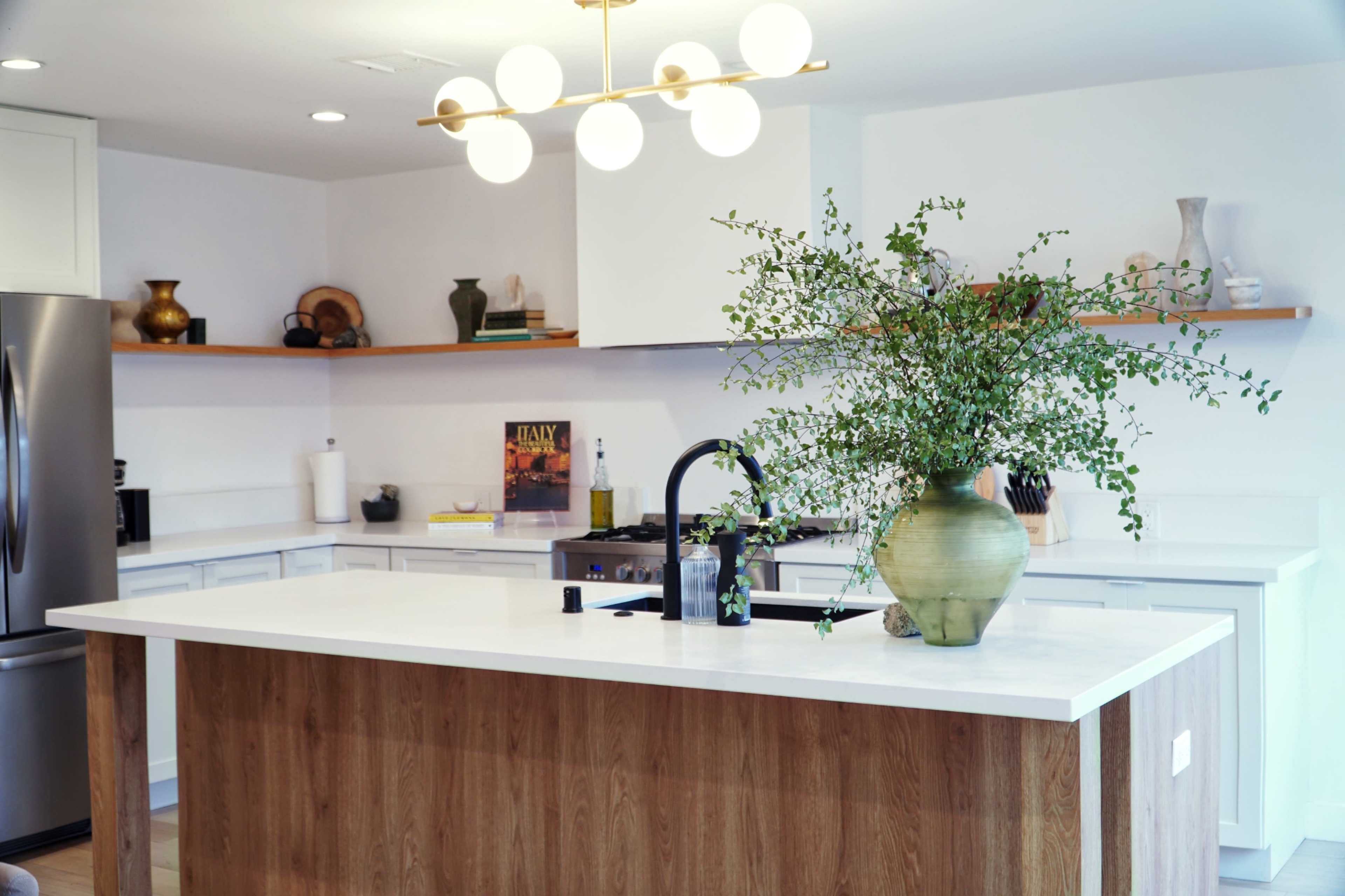 A modern kitchen features a large island with a light wood base, a black faucet, and a glass vase filled with greenery.