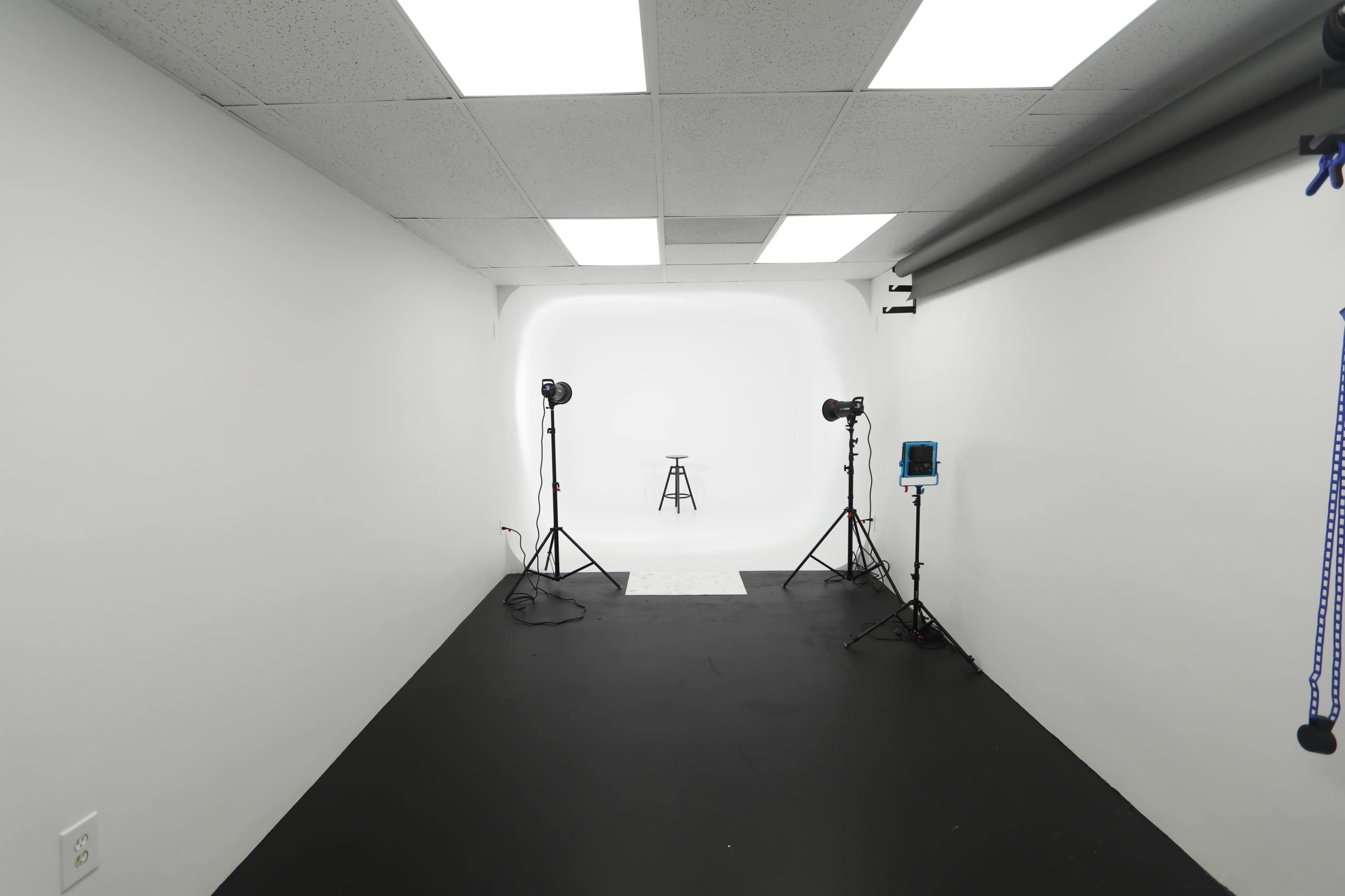 A clean, empty photography studio features a black floor, white walls, and a solitary stool illuminated by softbox lights on tripods.