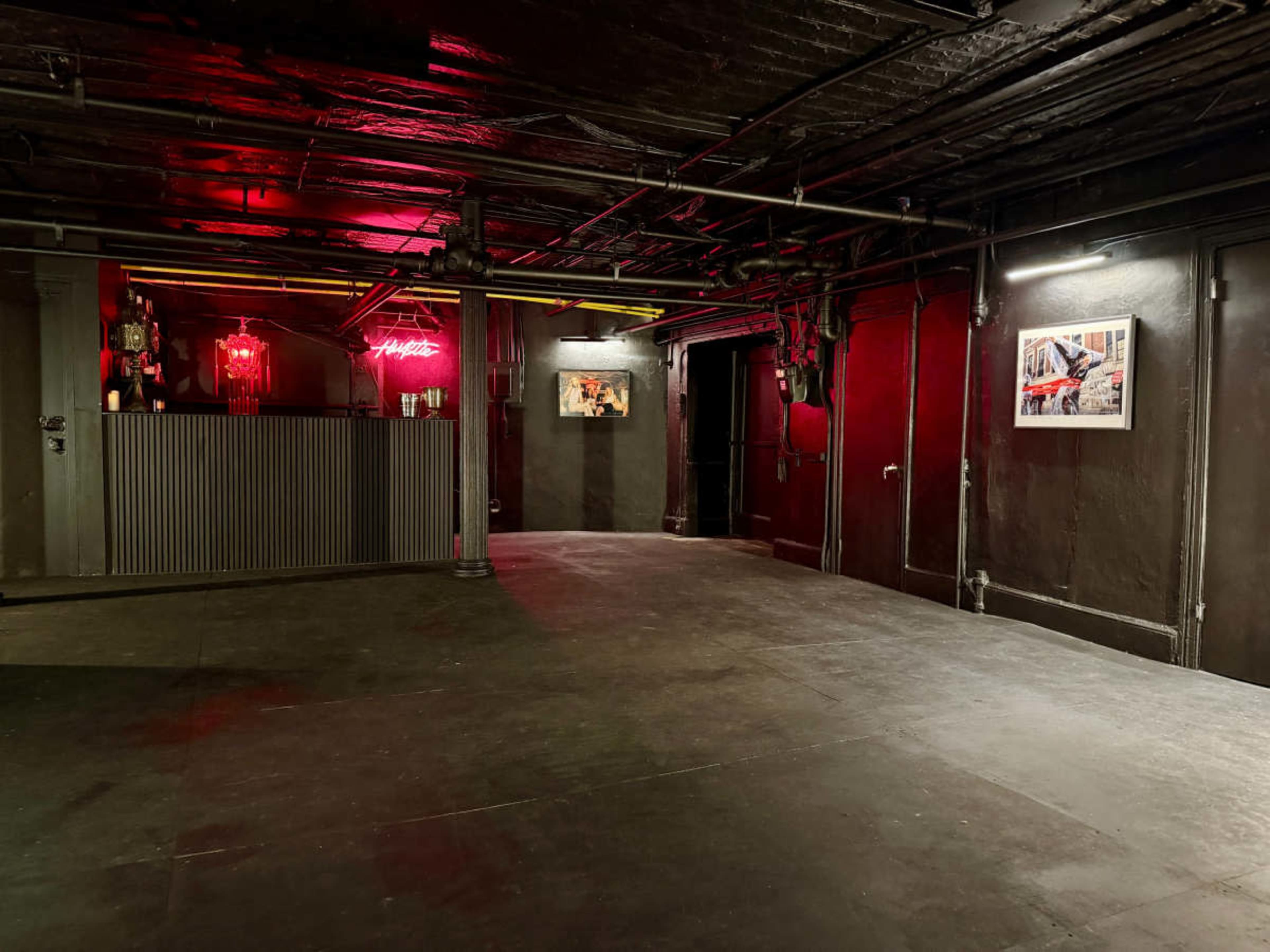 The image shows a dimly lit, empty underground space with red lighting, featuring a bar area and framed artworks on the walls.