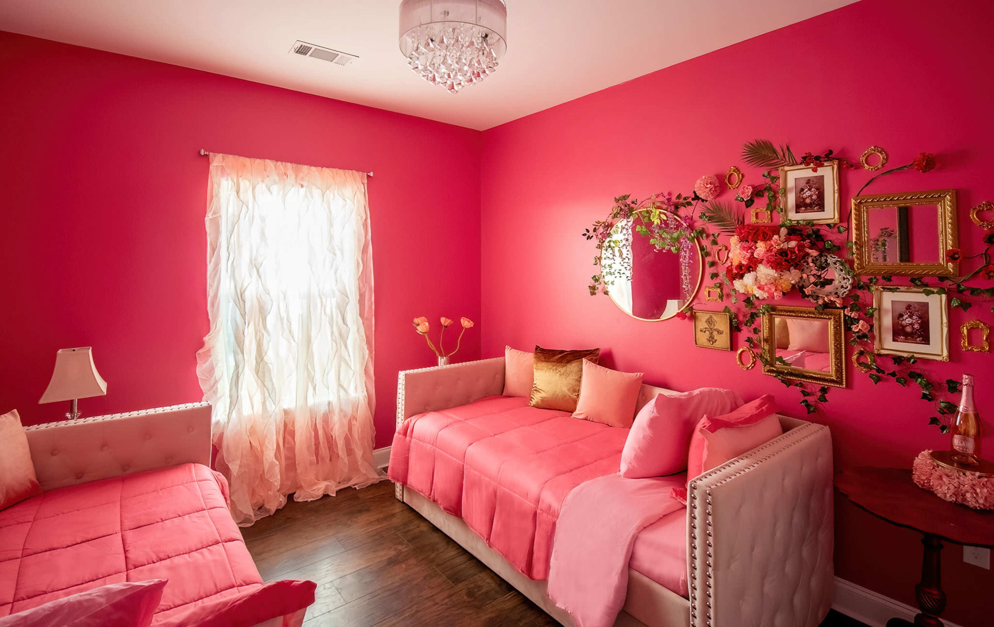 The image shows a pink-themed bedroom featuring two daybeds, a decorative mirror, and floral wall art.