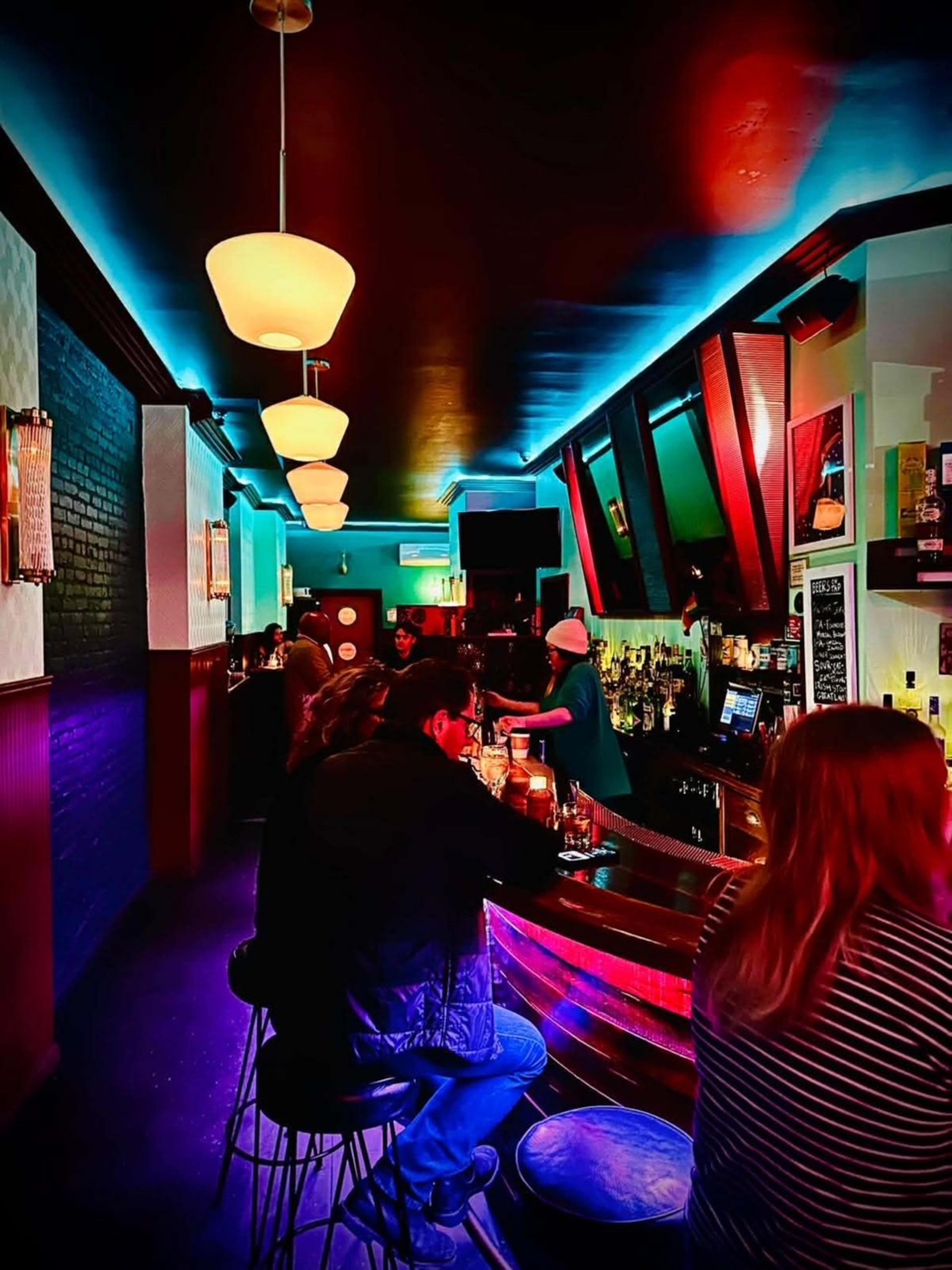 The image shows a dimly lit bar with patrons seated at the counter, illuminated by colorful overhead lighting.