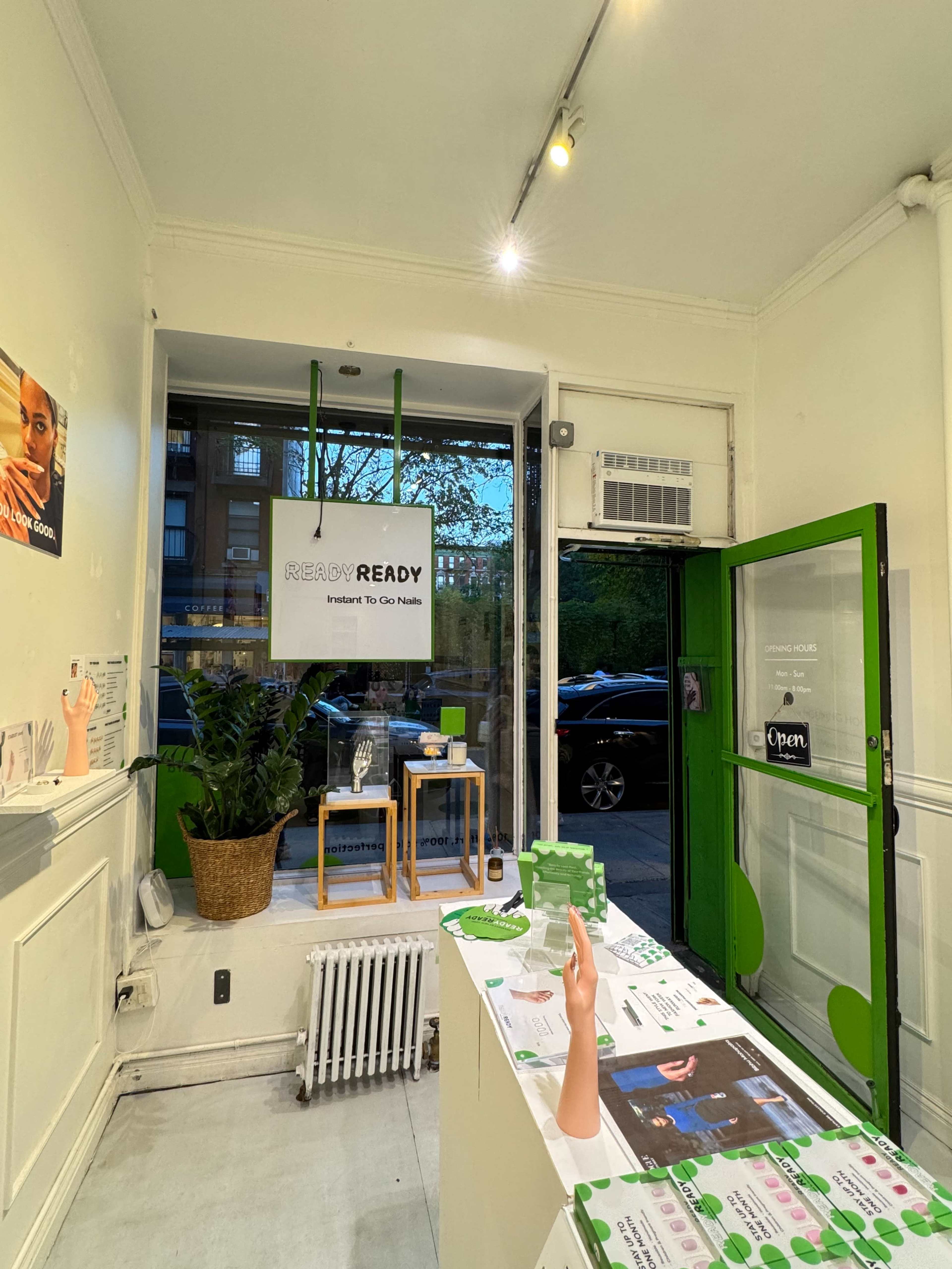 The interior of a bright, modern retail space features a white counter with display materials, a sign hanging from the ceiling, and a large window showing greenery outside.